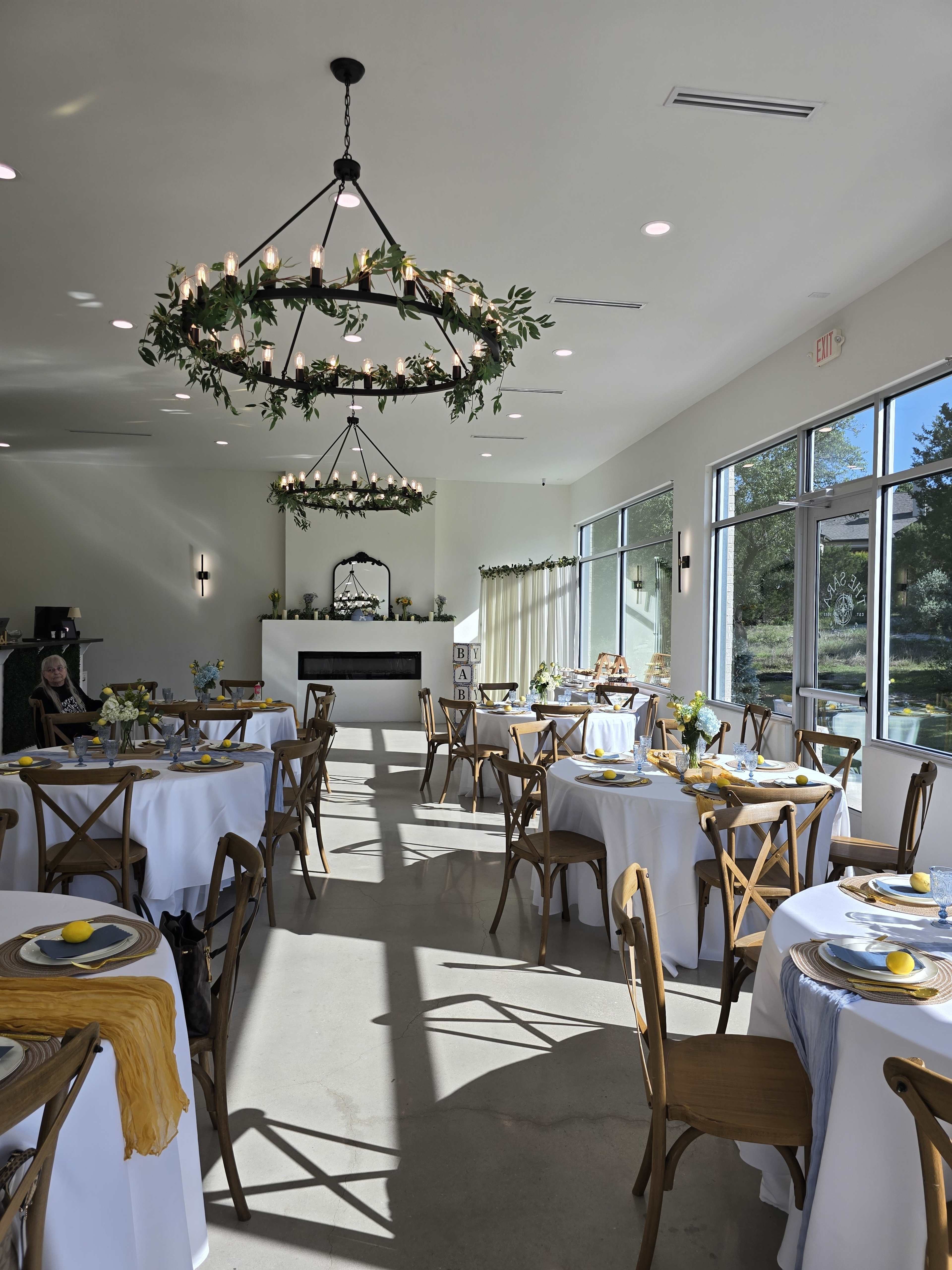 The image shows a bright, modern dining room with round tables set for an event, featuring floral centerpieces and decorative yellow accents.