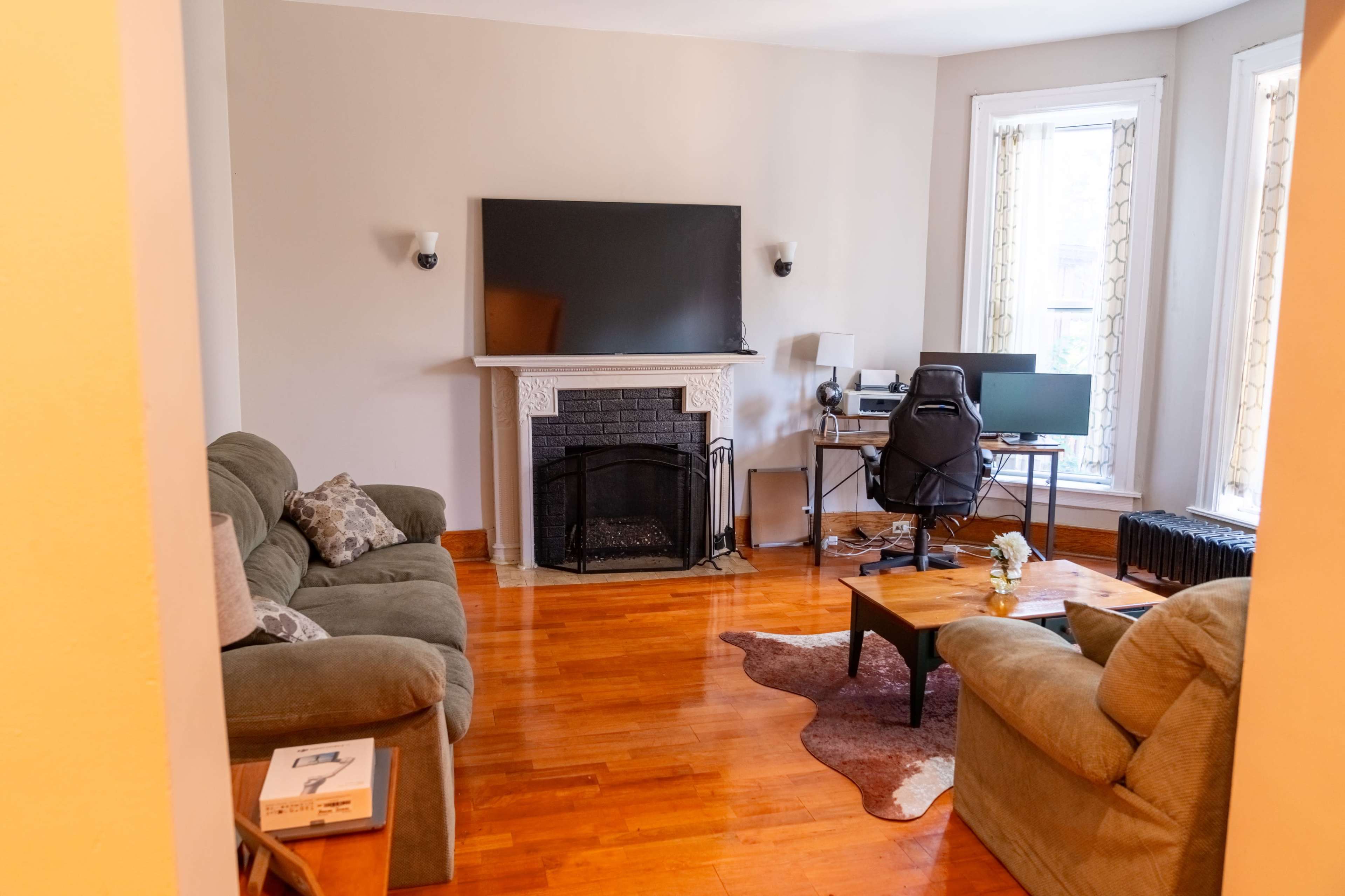 A living room with a television mounted above a fireplace, two couches, a coffee table, and a computer setup in a corner.