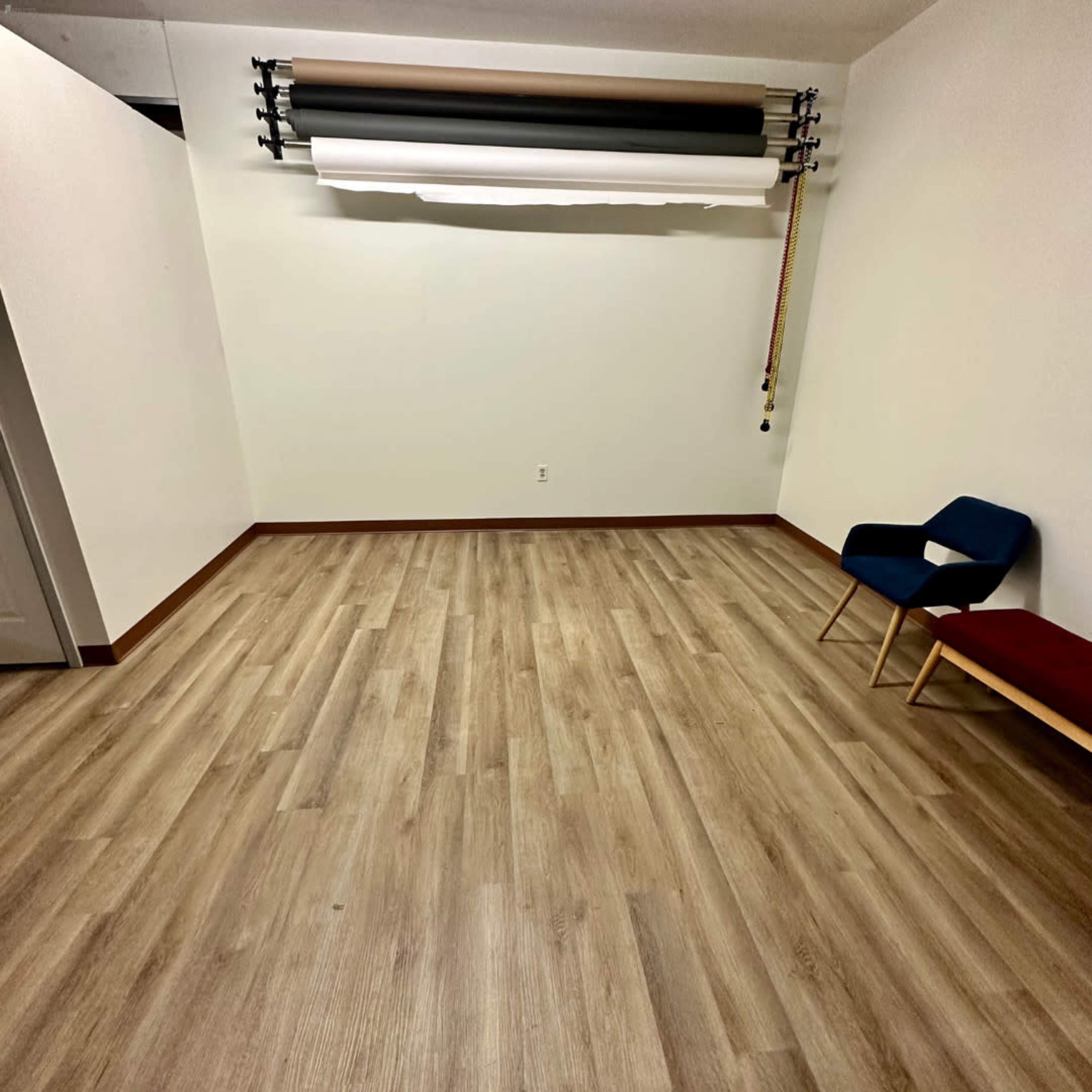 The image shows a sparsely furnished room with light wood flooring, white walls, and a blue chair positioned next to a red bench.