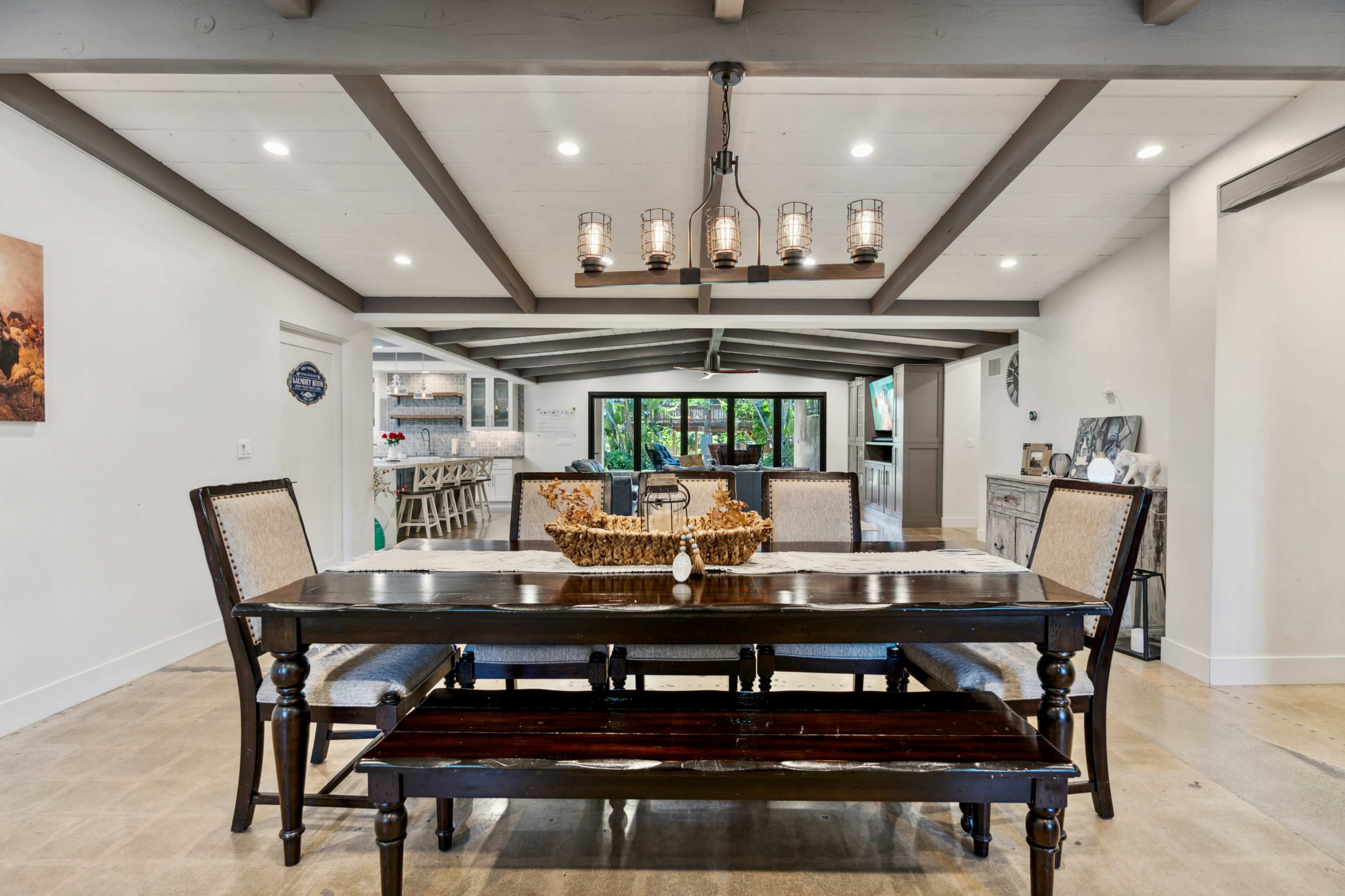 A dining room features a wooden table with a bench and chairs beneath a chandelier, surrounded by white walls and a beamed ceiling.