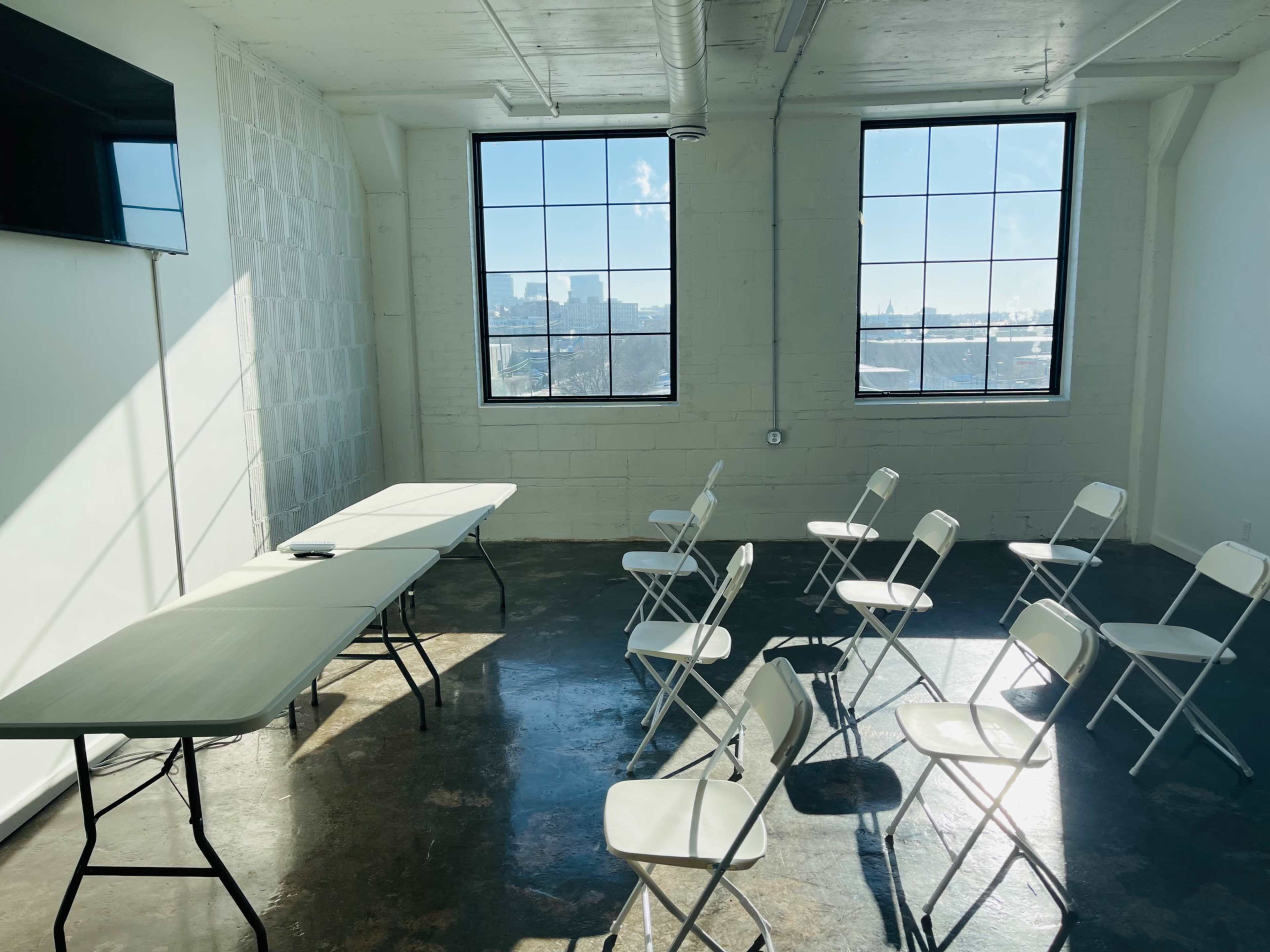 A small meeting room features two tables at the front and several white folding chairs arranged in a semicircle facing a large window.
