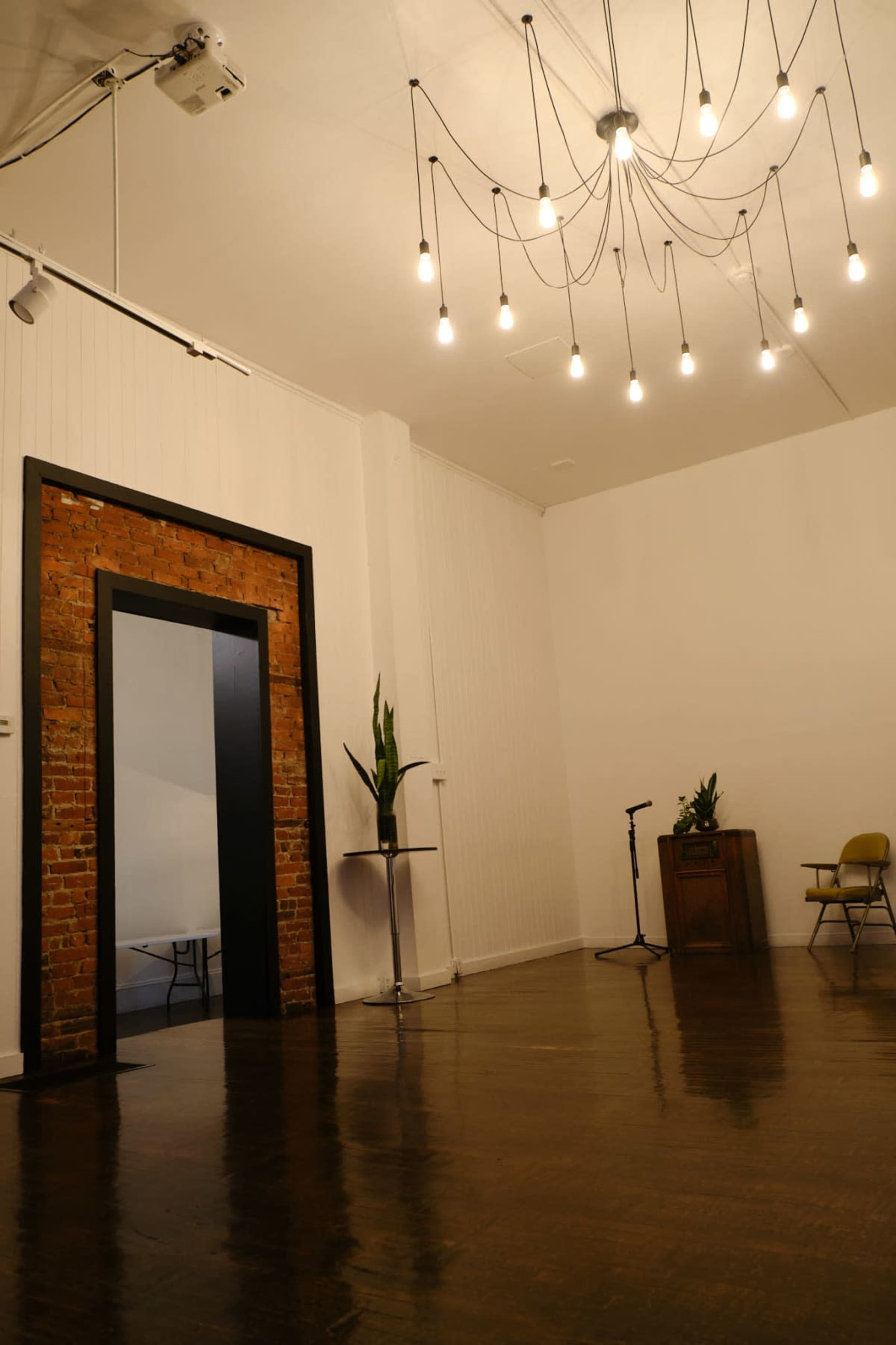 The image shows a spacious room with wooden flooring, a large brick archway, a chandelier of hanging light bulbs, and minimal furnishings including a microphone, a plant, and a chair.