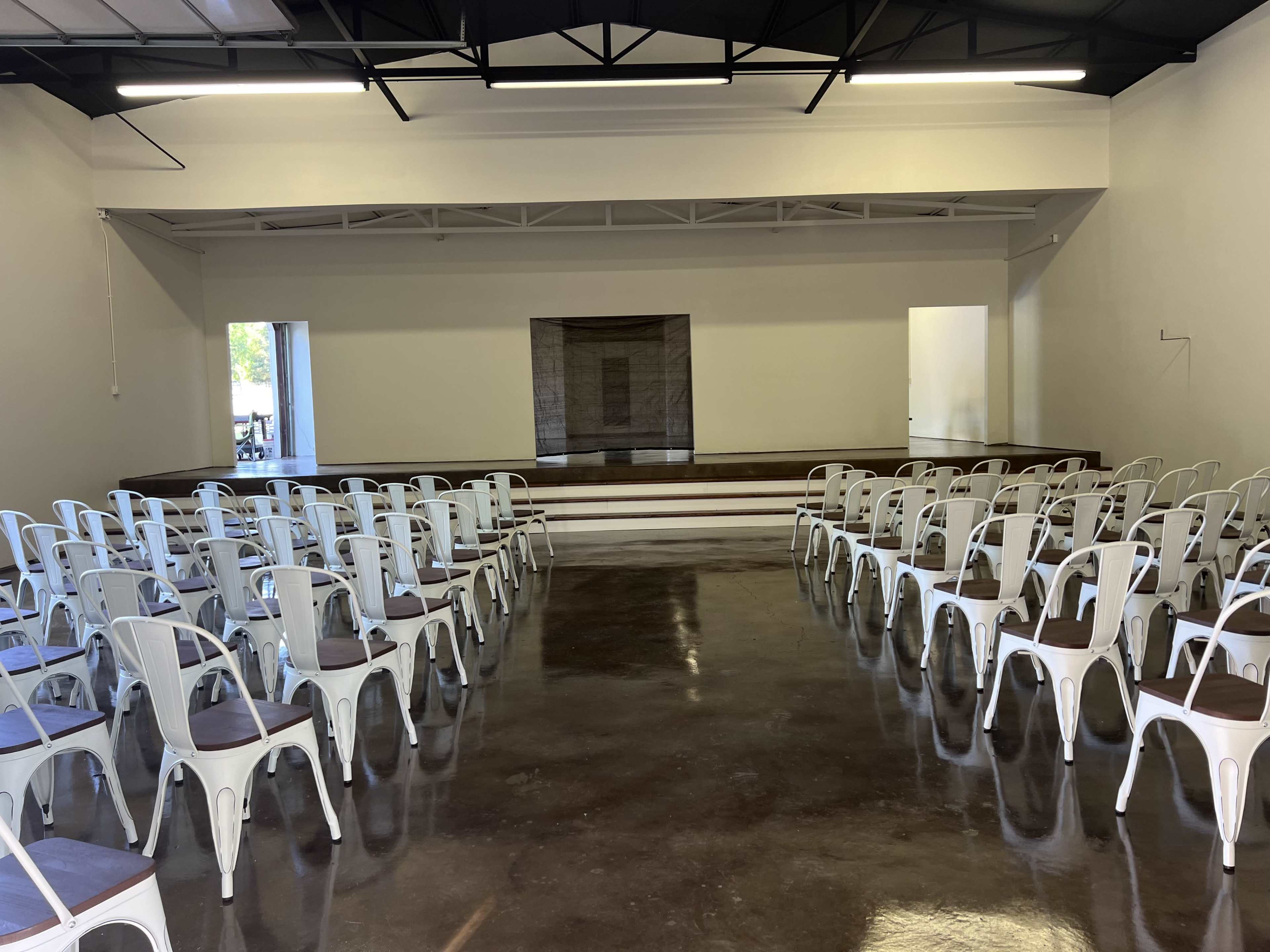 The image shows a spacious room with metal chairs arranged in rows facing a stage at the front.