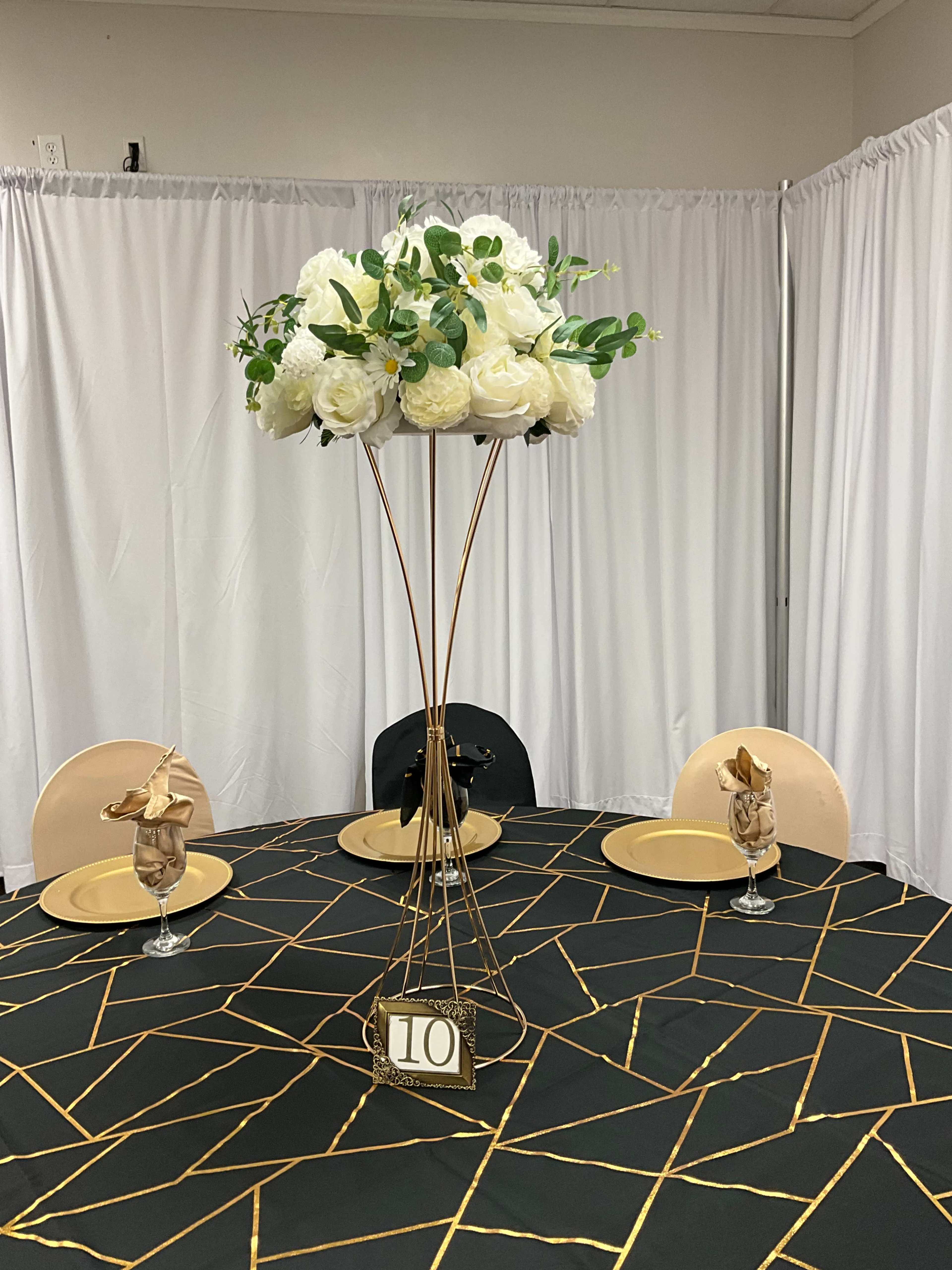 A round dining table is set with a geometric black and gold tablecloth, featuring a tall floral centerpiece and elegantly arranged place settings.