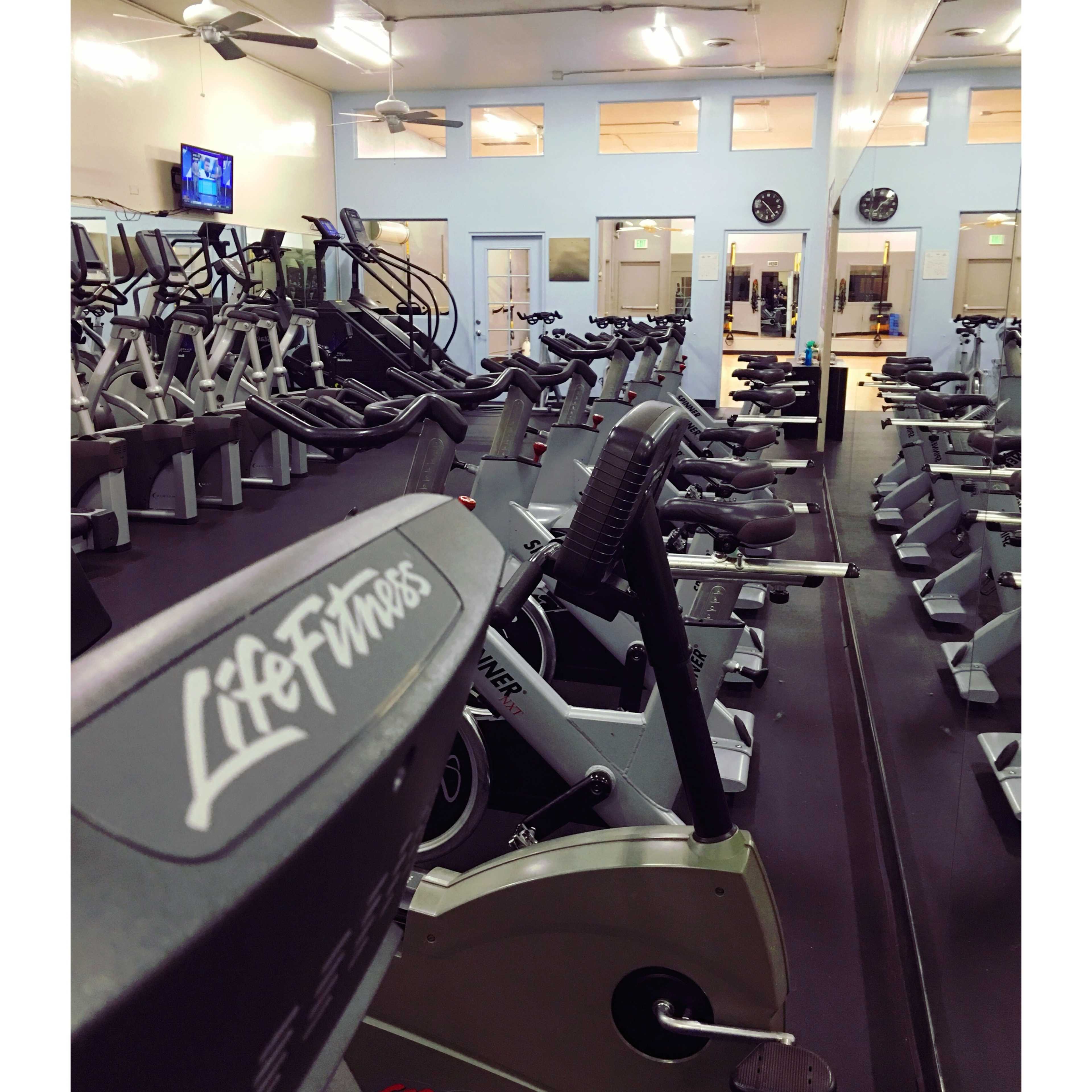 A row of exercise machines is lined up in a well-lit gym, with mirrors reflecting the equipment and wall clocks.