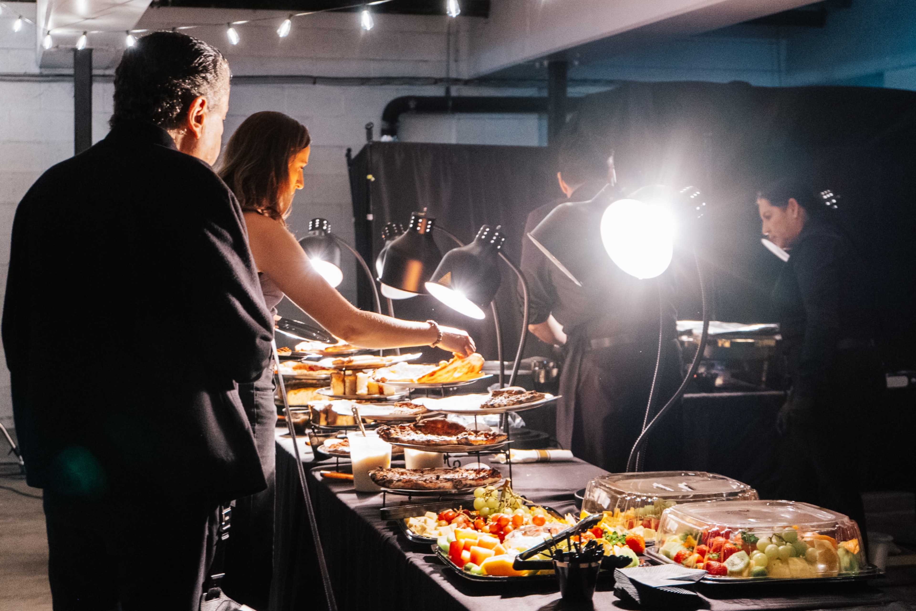A group of people is serving themselves food from a long table under bright lights at a catered event.