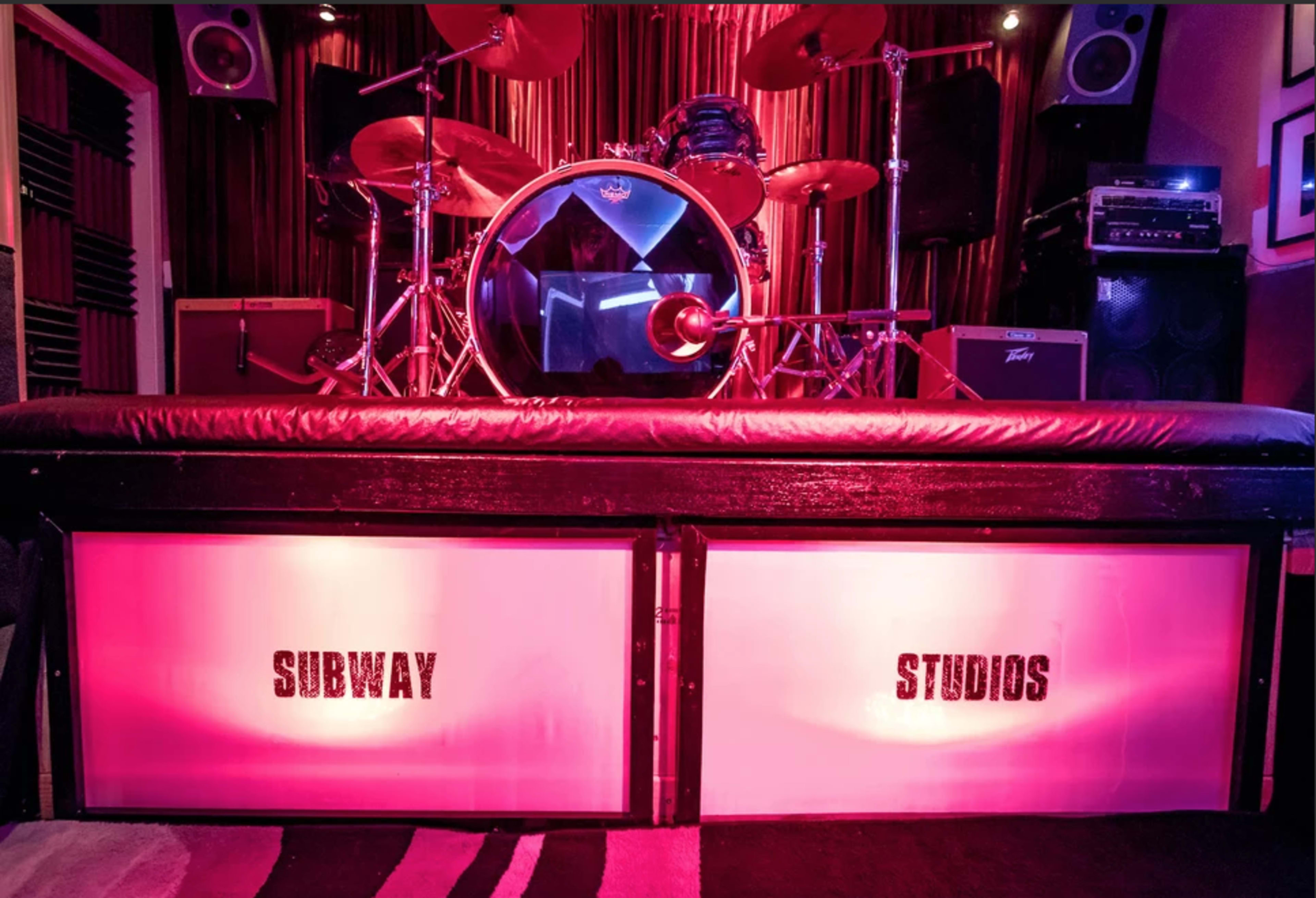 The image shows a music studio setup featuring a drum kit and two illuminated display cases labeled "SUBWAY" and "STUDIOS."