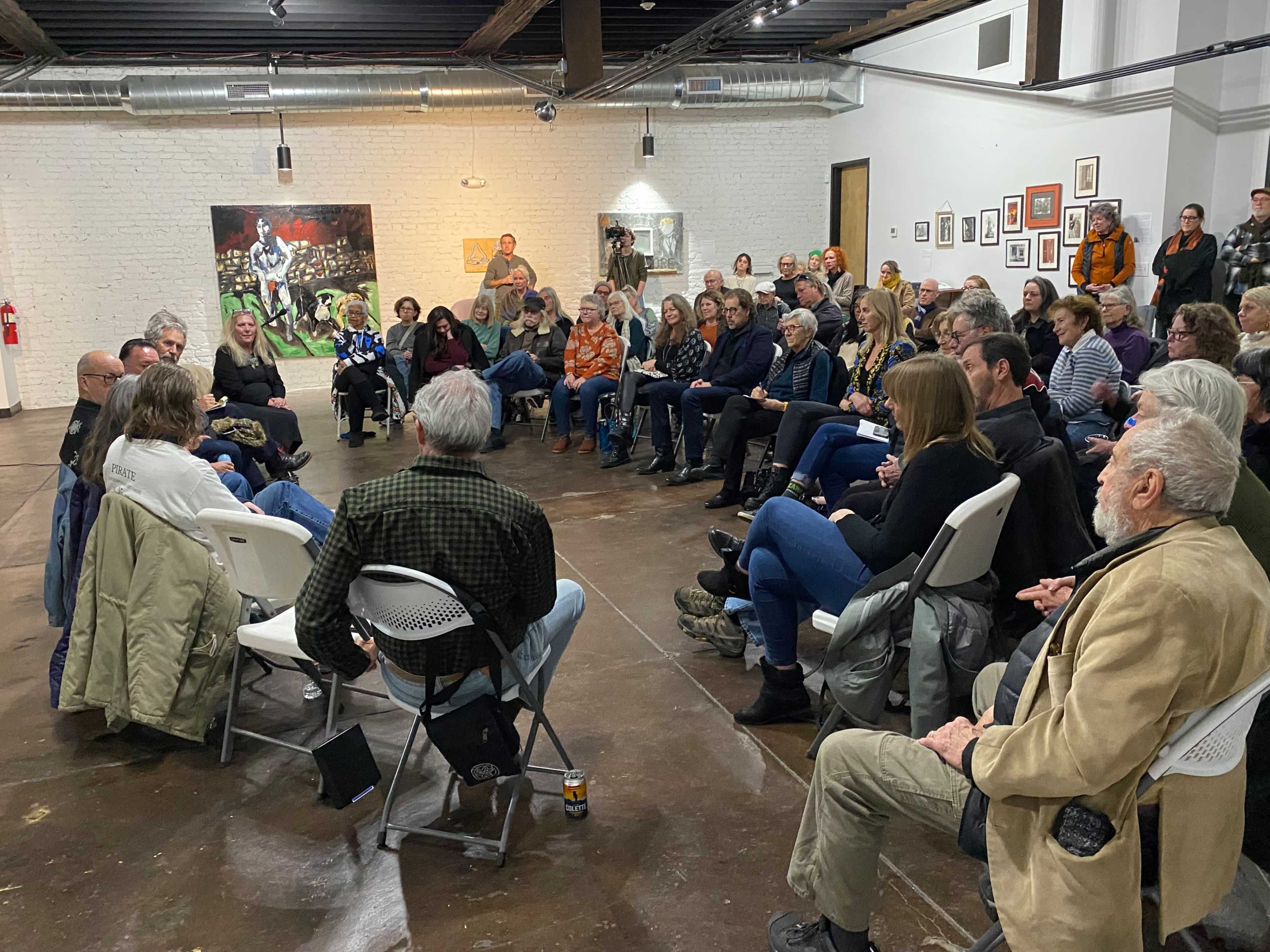 A large group of people sits in a gallery for a discussion event, with a panel of speakers at the front.
