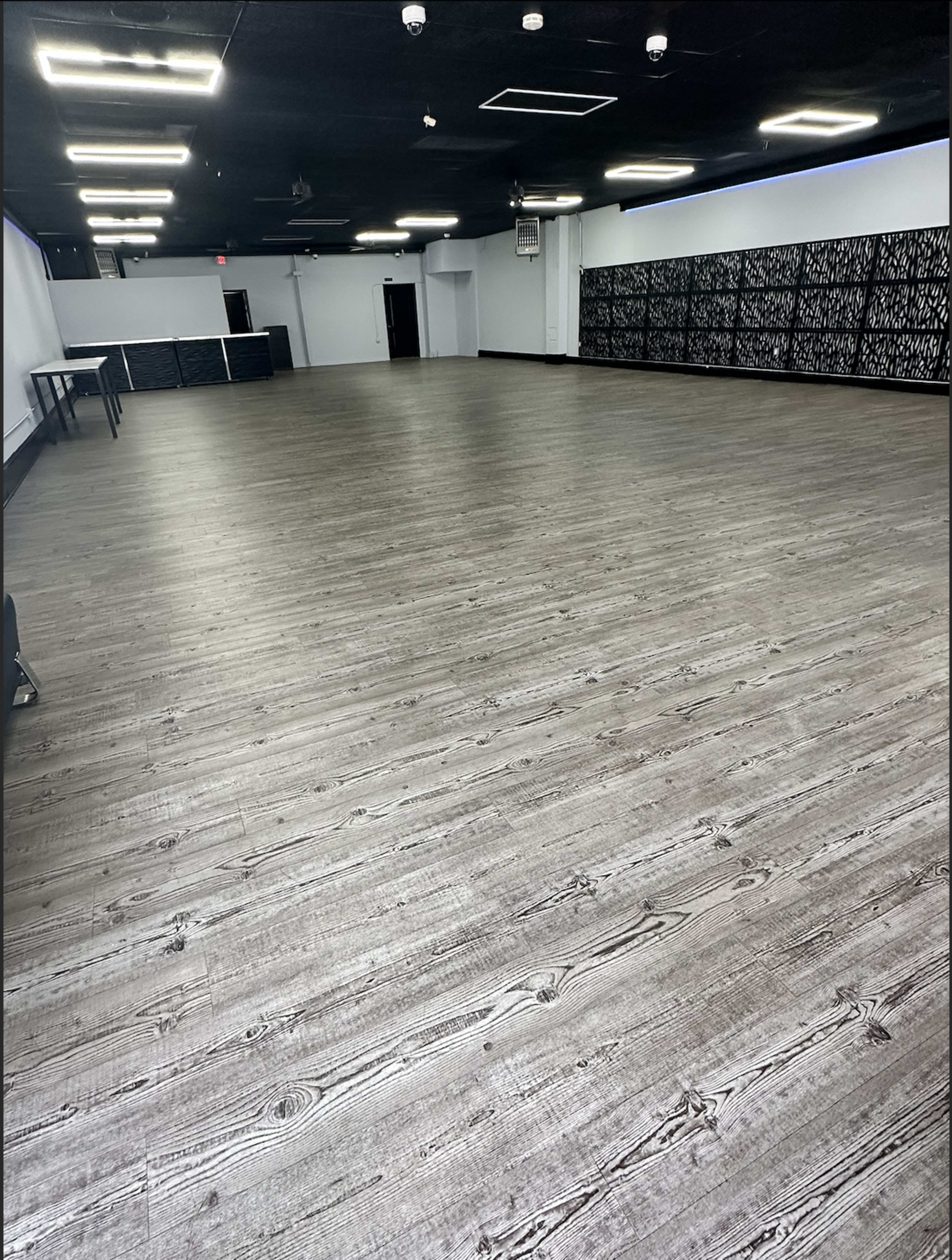The image shows a spacious empty room with wooden-style flooring and black walls in the background.