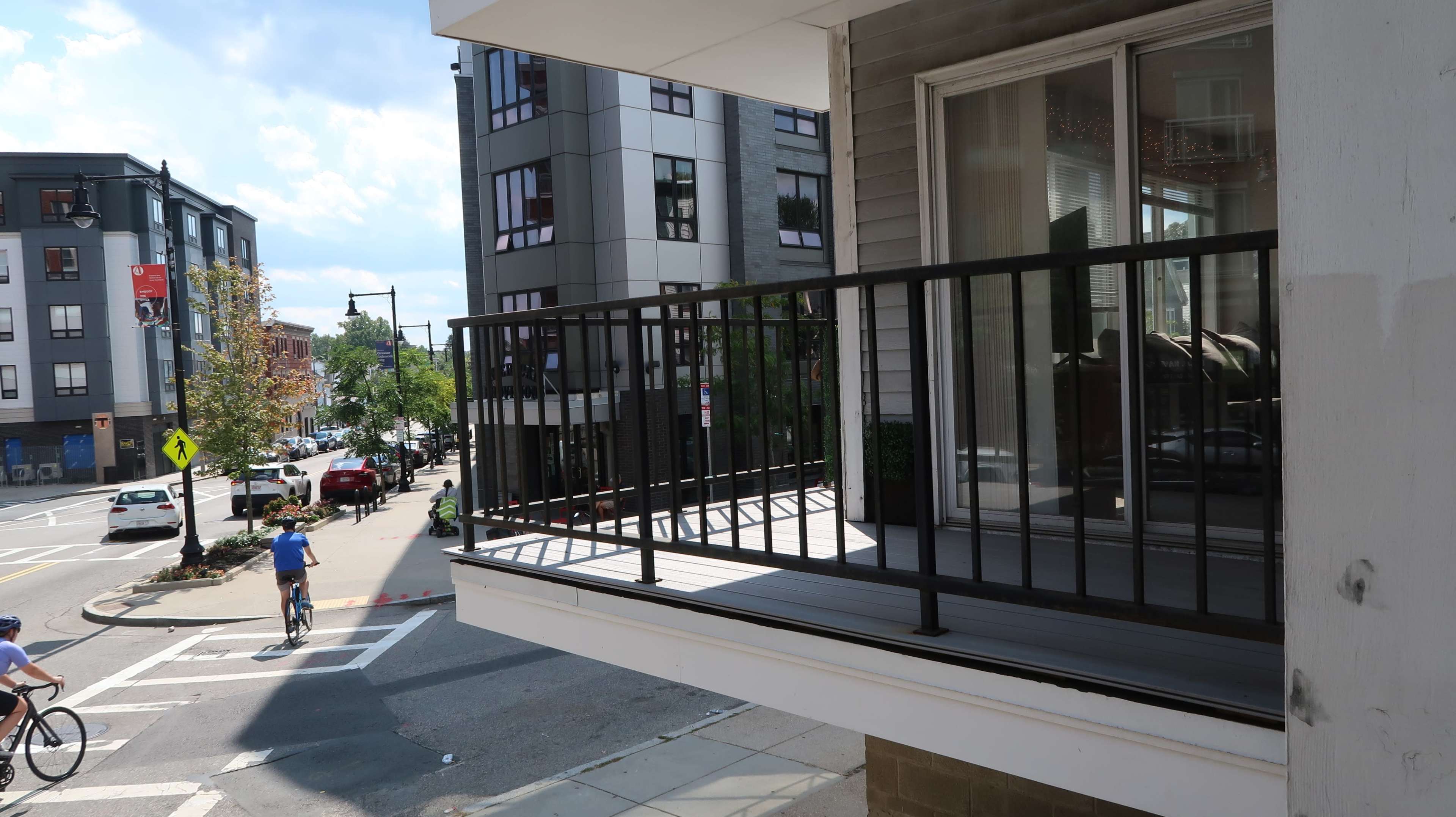A balcony with a black railing overlooks a street corner with parked cars and cyclists.