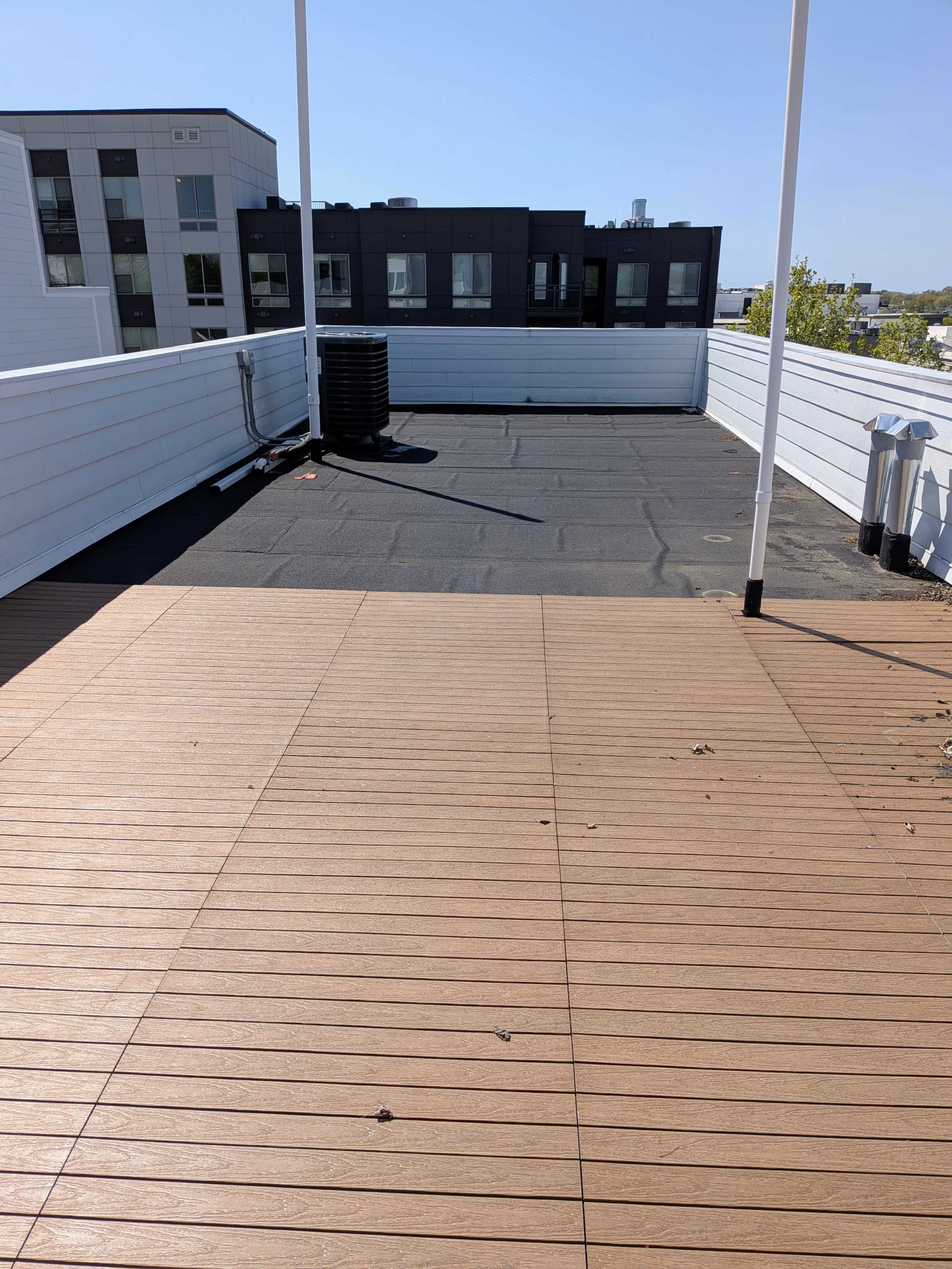 The image shows a rooftop terrace with a wooden deck on one side and a black flat surface on the other, surrounded by white railings and buildings in the background.