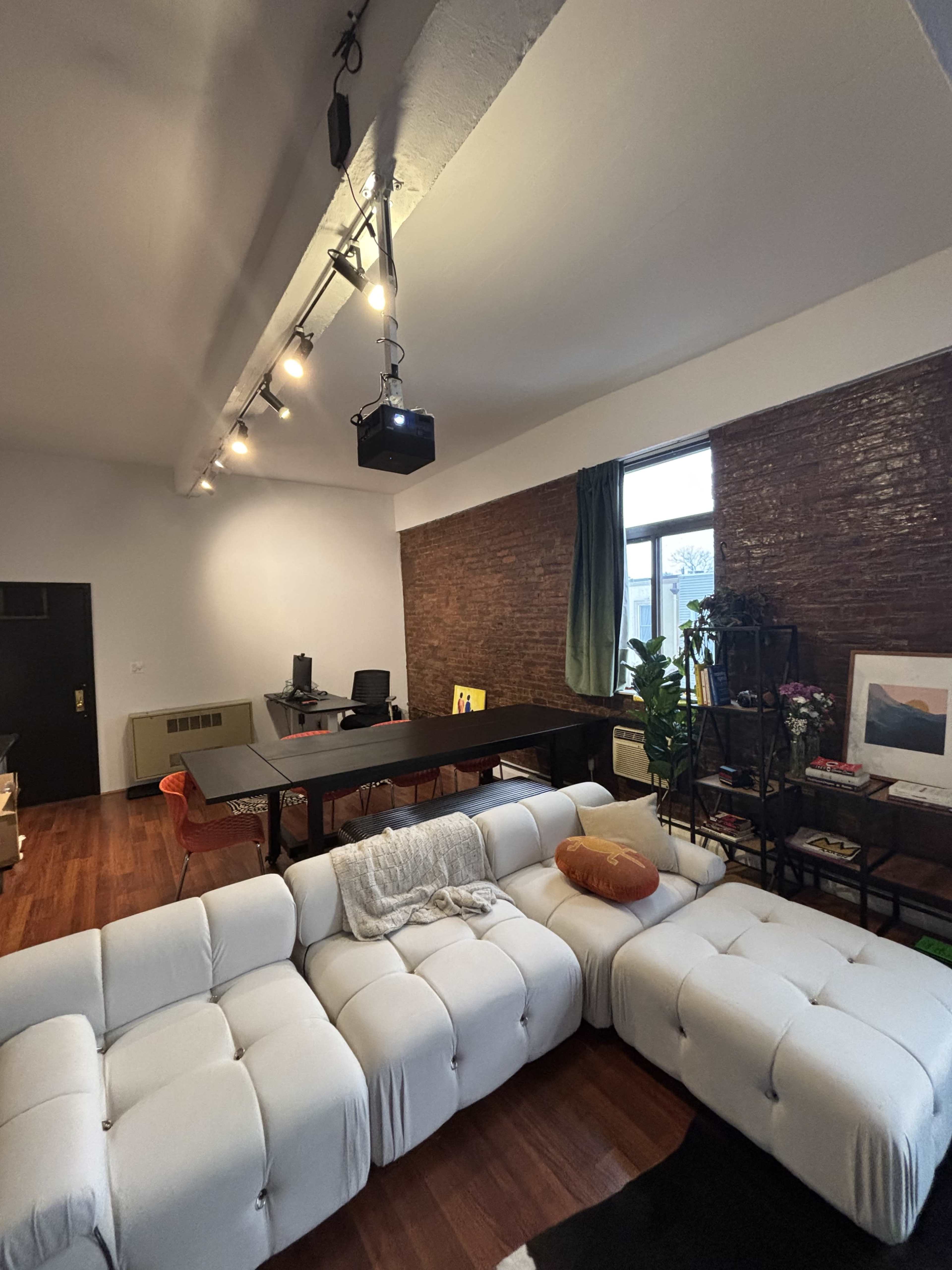 The image shows a modern living space featuring a white sectional couch, a dark wooden floor, a large window, and a workspace with a desk and chairs in the background.