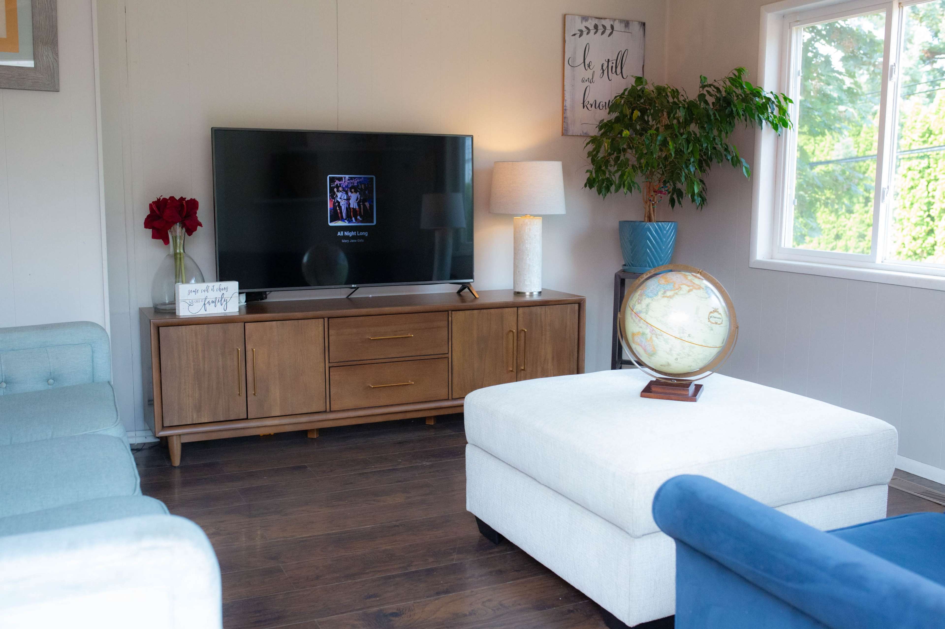 The living room features a television on a wooden cabinet, flanked by a lamp and a globe on an ottoman, with a plant and decorative flowers in the background.