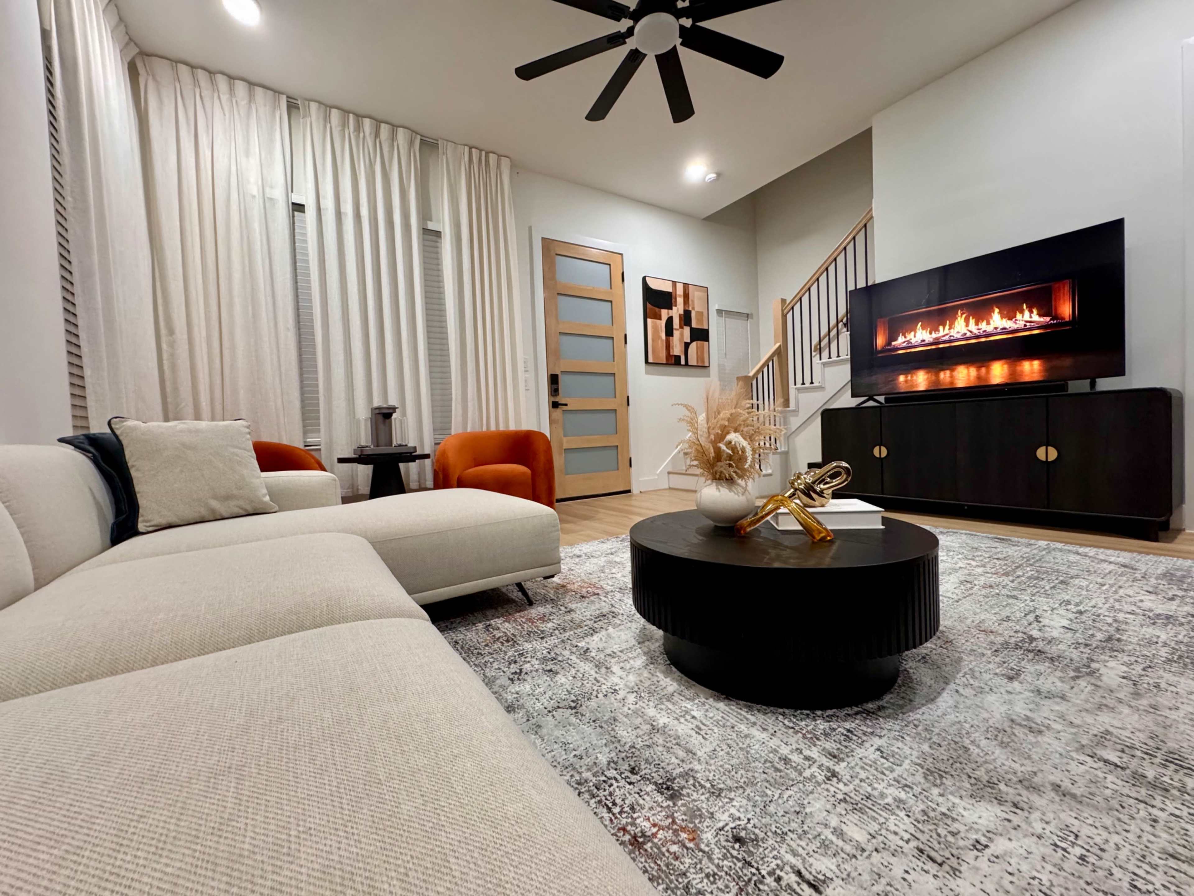 The image shows a modern living room featuring a sectional sofa, orange accent chairs, a coffee table, and a wall-mounted electric fireplace.