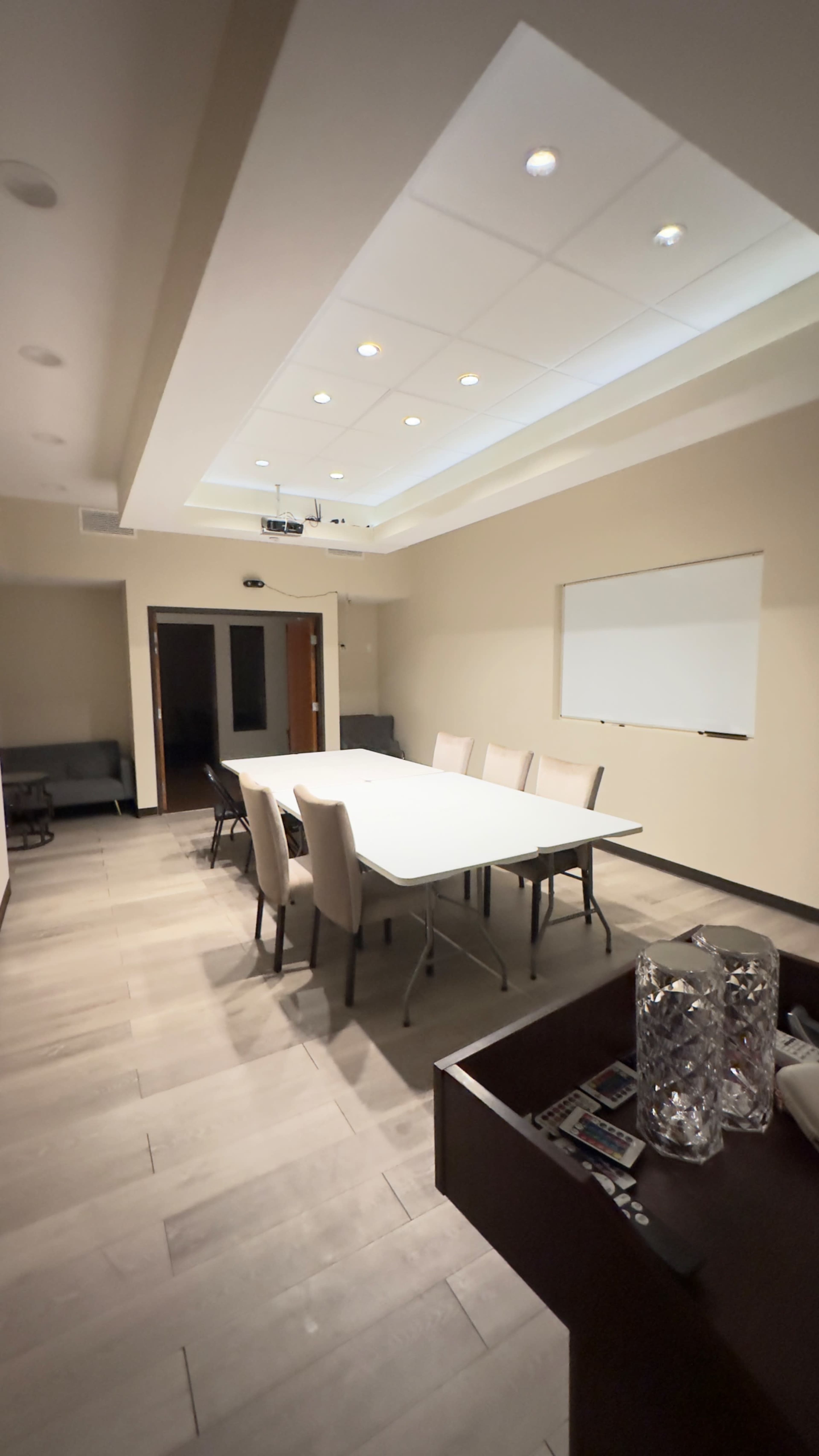 The image shows a conference room with a long table surrounded by chairs, a whiteboard on the wall, and a small seating area in the background.