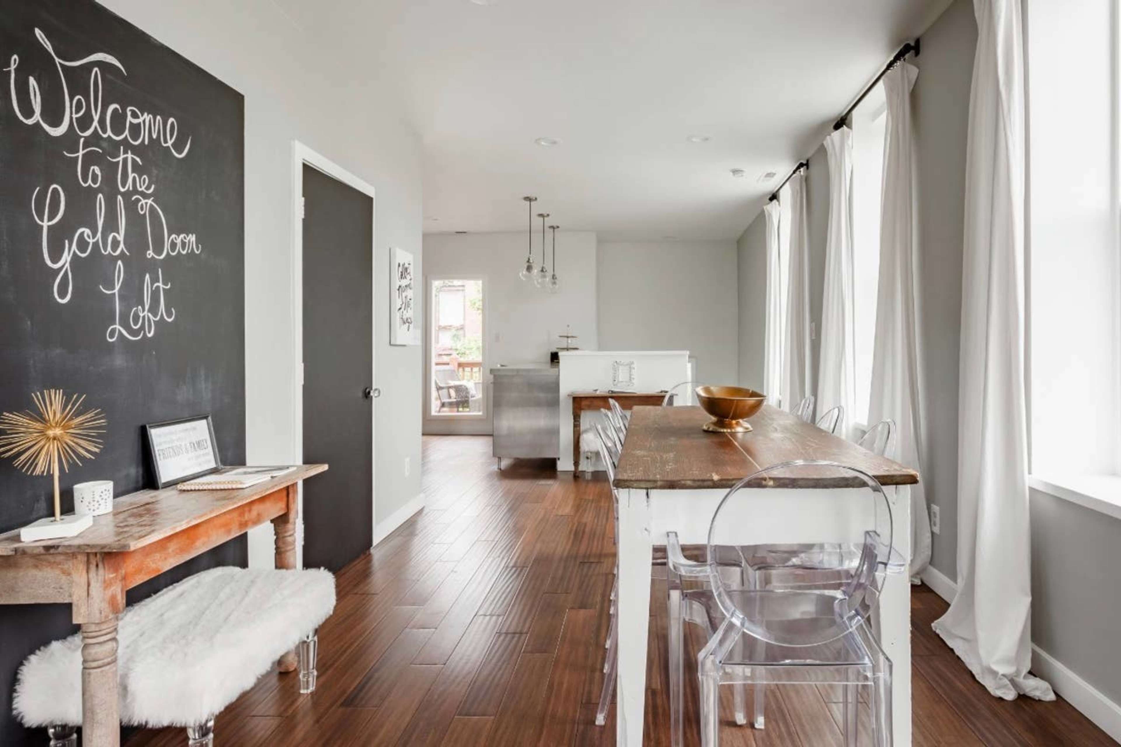 A modern dining area featuring a wooden table, transparent chairs, a chalkboard wall with a welcome message, and large windows with white curtains.