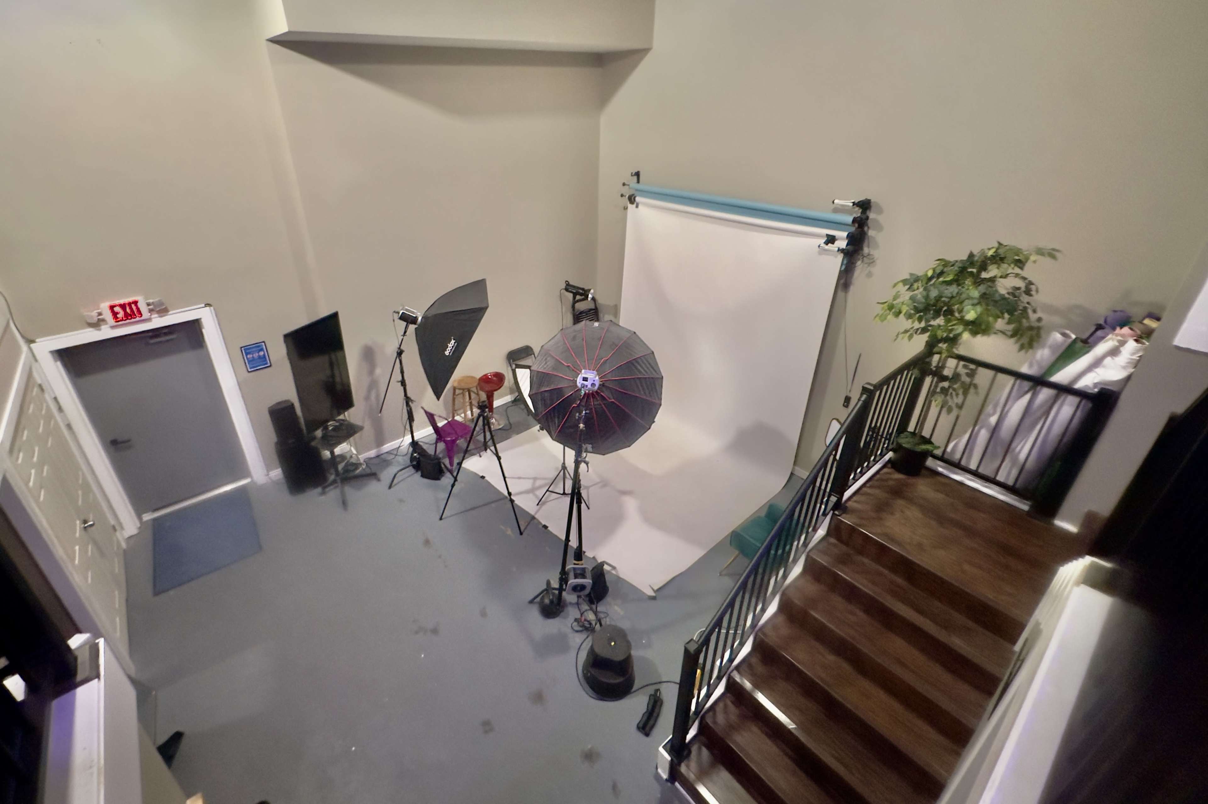 The image shows a photography studio with lighting equipment and a backdrop, along with a staircase and a potted plant.