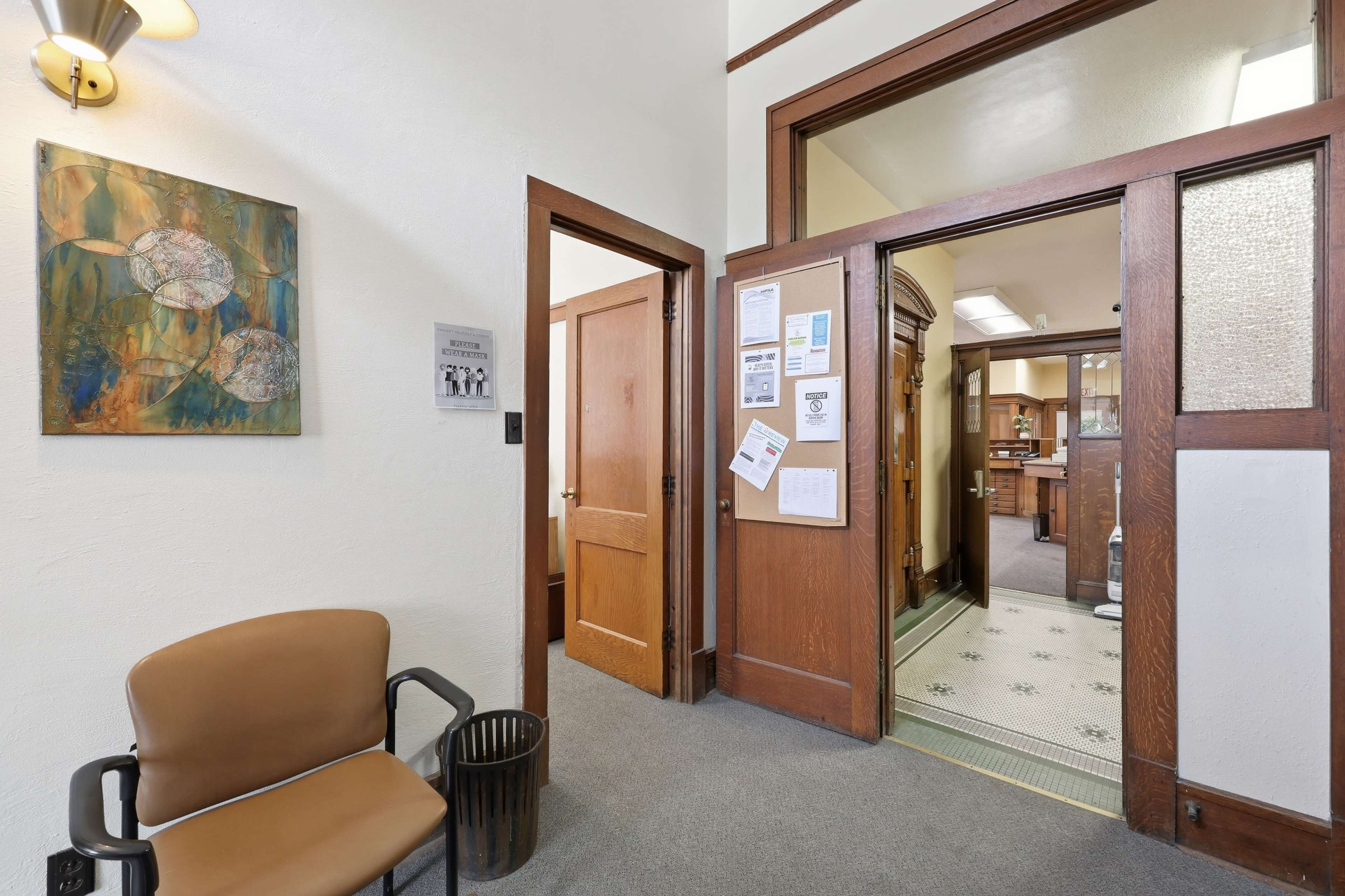 The image shows a reception area with a chair, a wastebasket, and a doorway leading into another room decorated with wooden features.