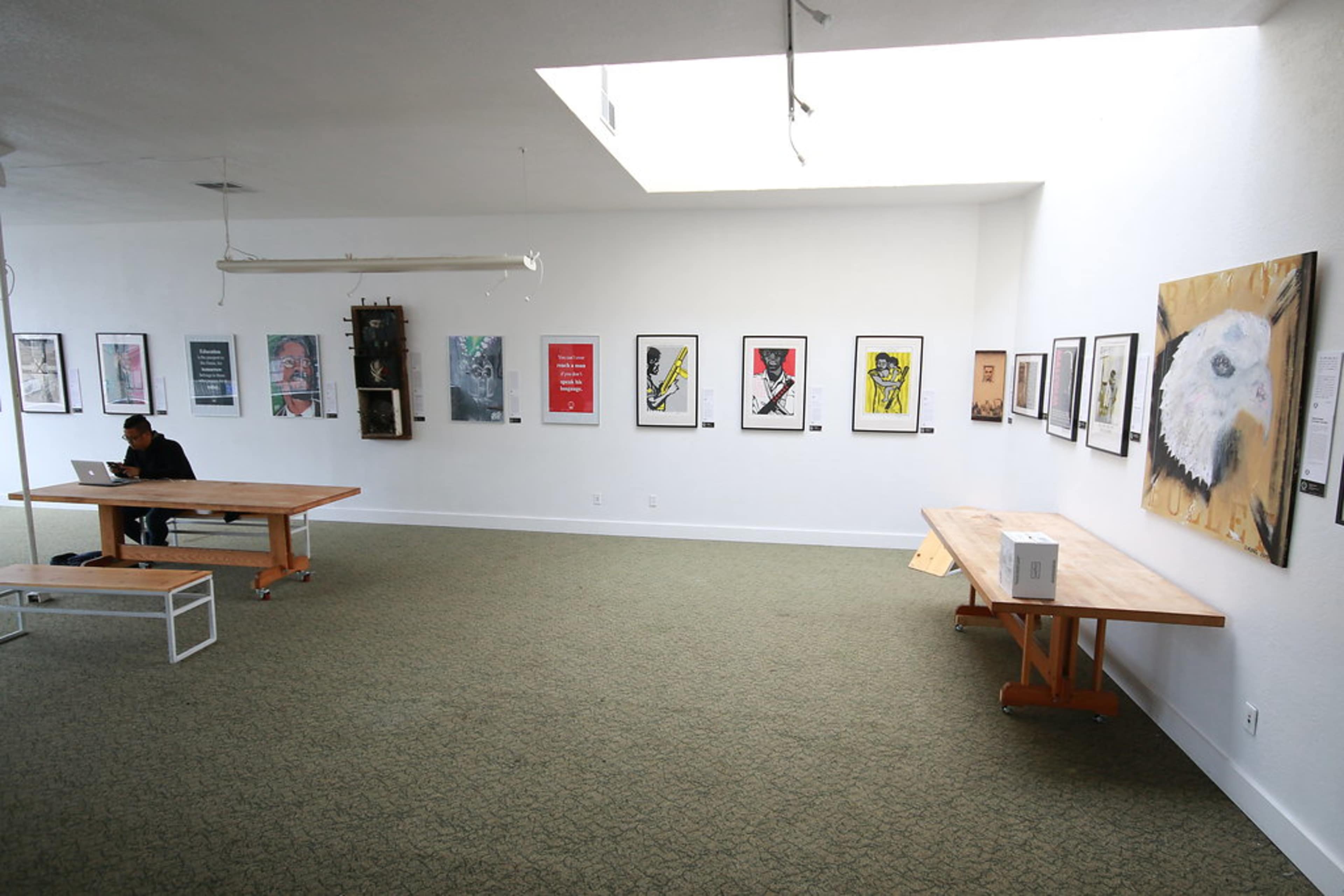 An art gallery with a person seated at a table while various framed artworks hang on the white walls.