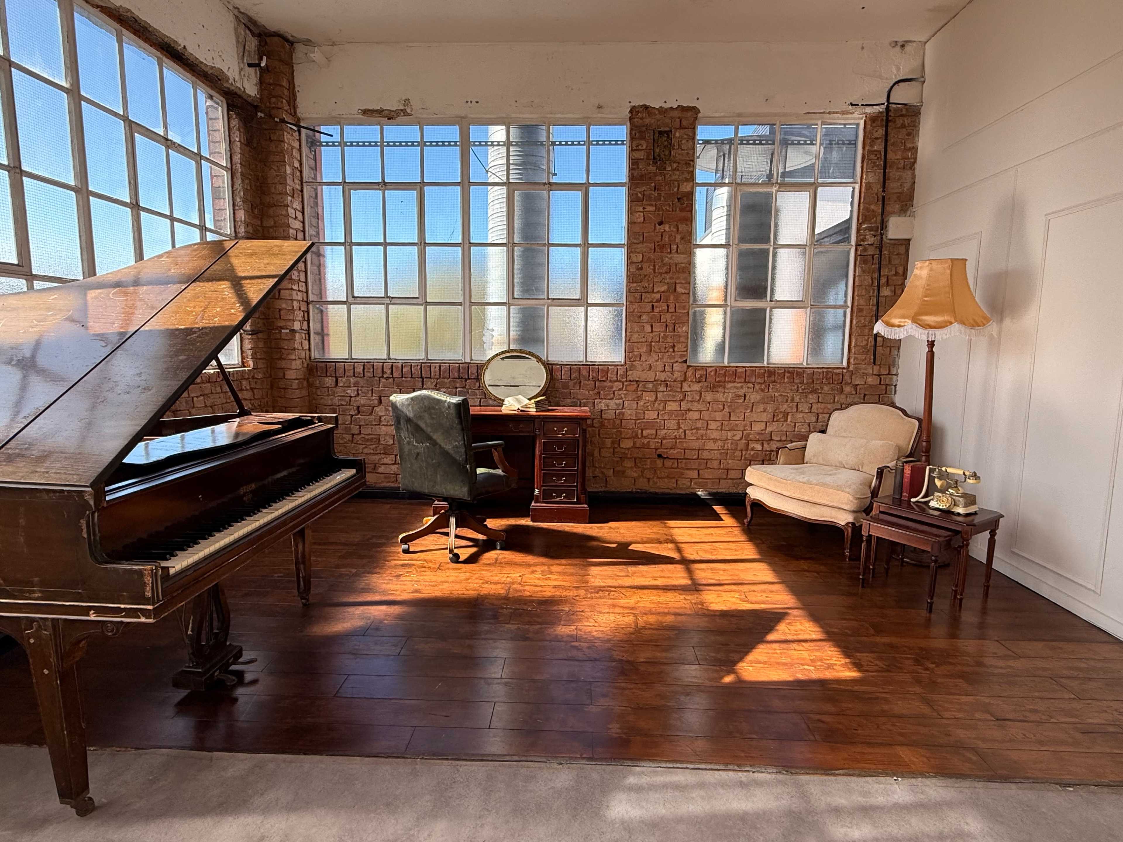 The image features a spacious room with large windows, a grand piano, a desk with a mirror, a chair, and a side table with a lamp.