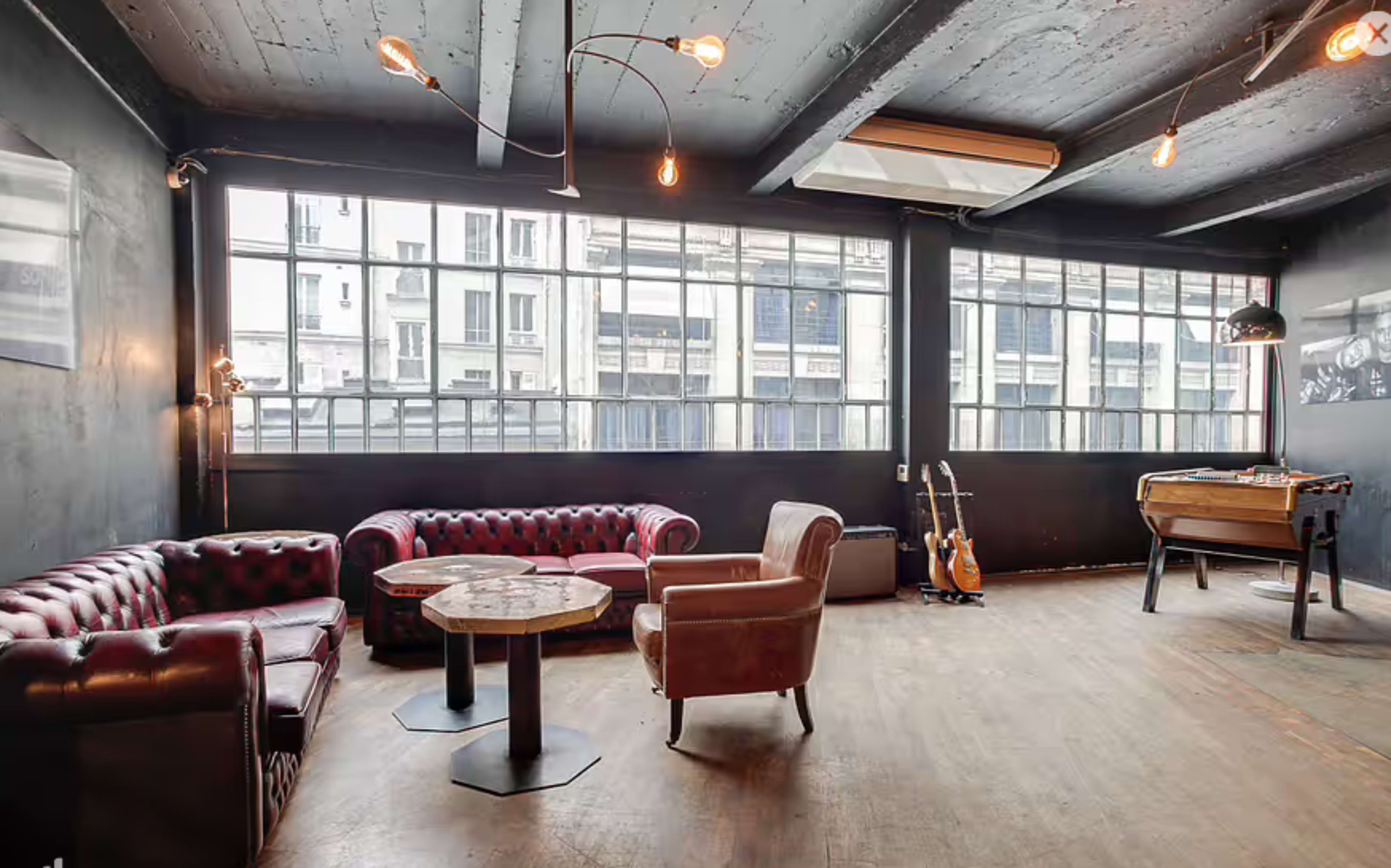 The image shows a spacious lounge area featuring leather sofas, a wooden table, and a foosball table, with large windows allowing natural light to enter.