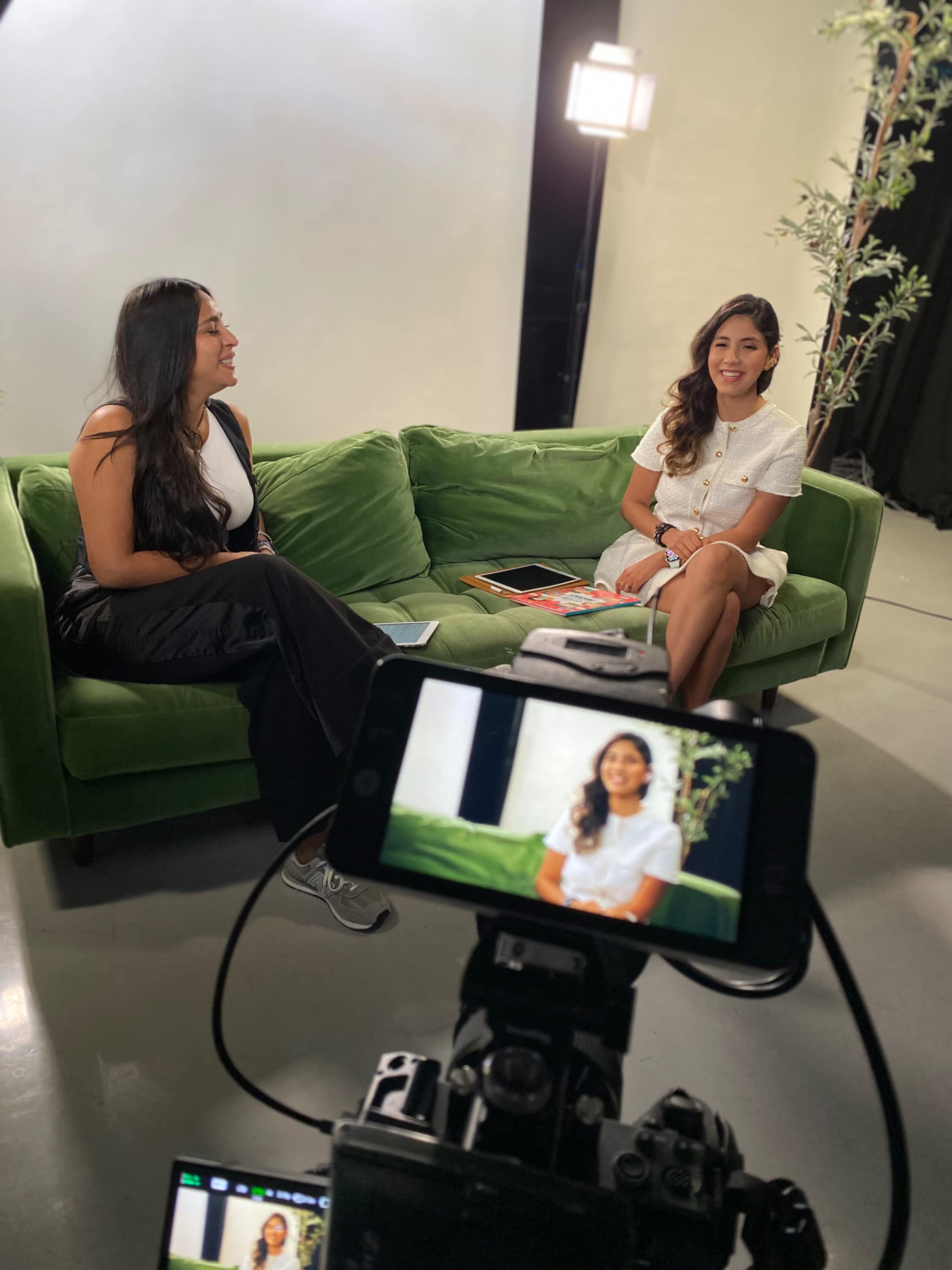 Two women are seated on a green couch, engaged in a conversation, while a camera captures the scene.