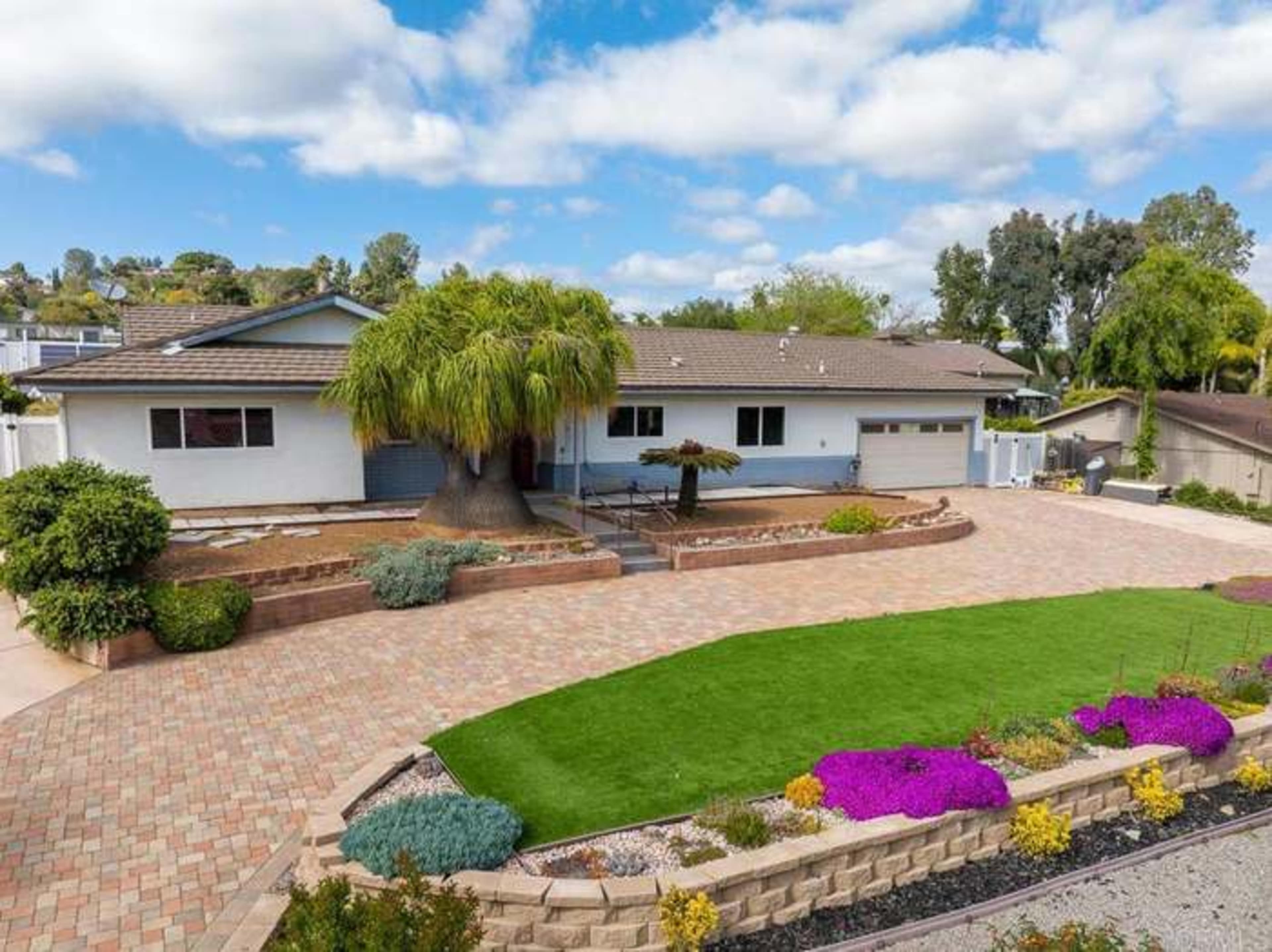 The image shows a single-story house with a landscaped front yard featuring a central tree and flower beds, surrounded by a brick driveway.
