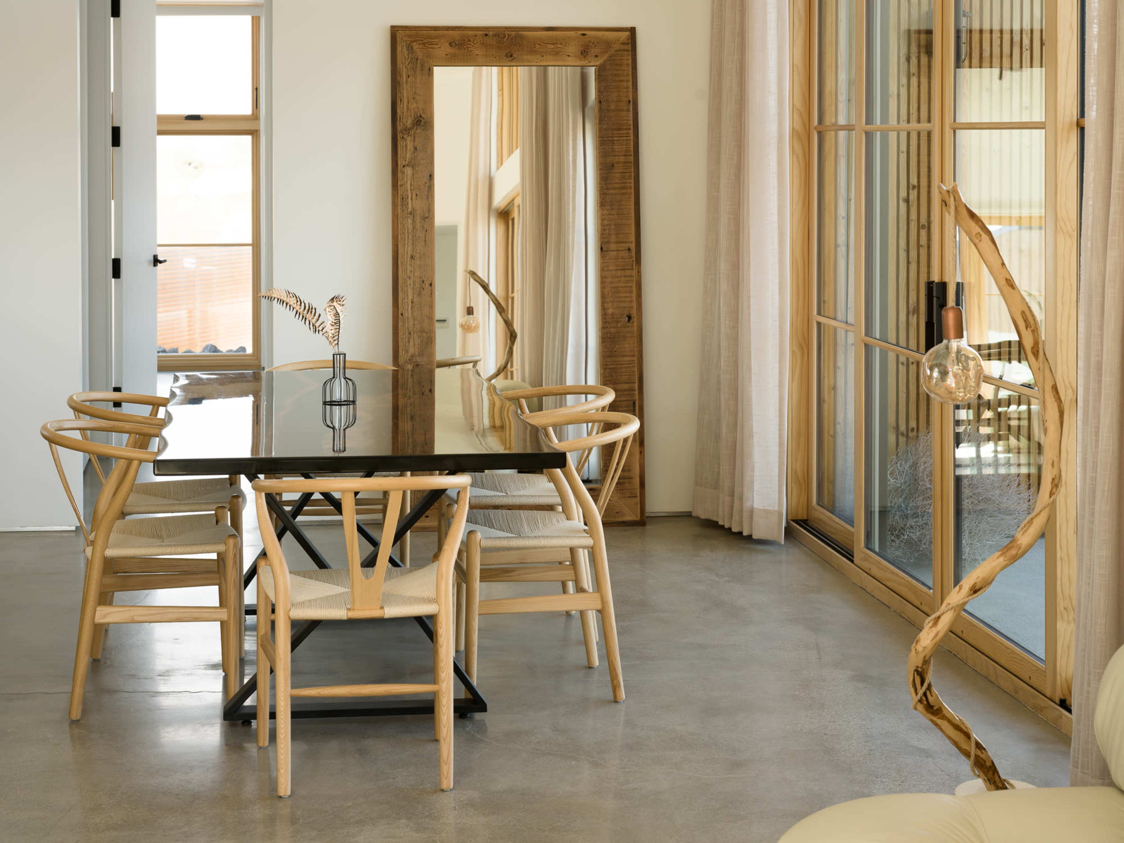 The image features a modern dining area with a large black table surrounded by six wooden chairs, a tall mirror, and a sliding glass door leading to an outdoor space.