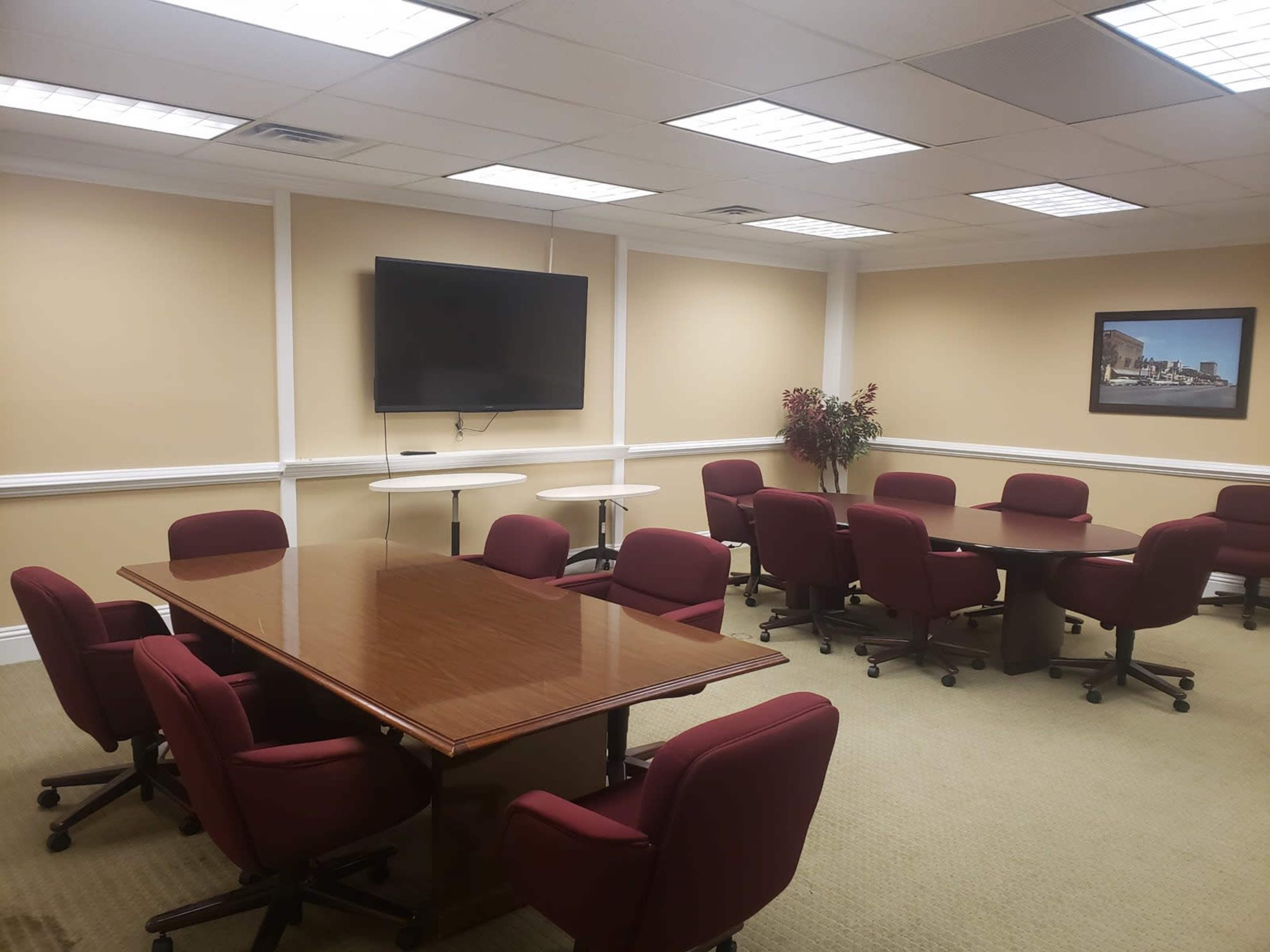 A conference room features several maroon chairs arranged around two tables and a wall-mounted television, with light-colored walls and a framed picture on the far wall.