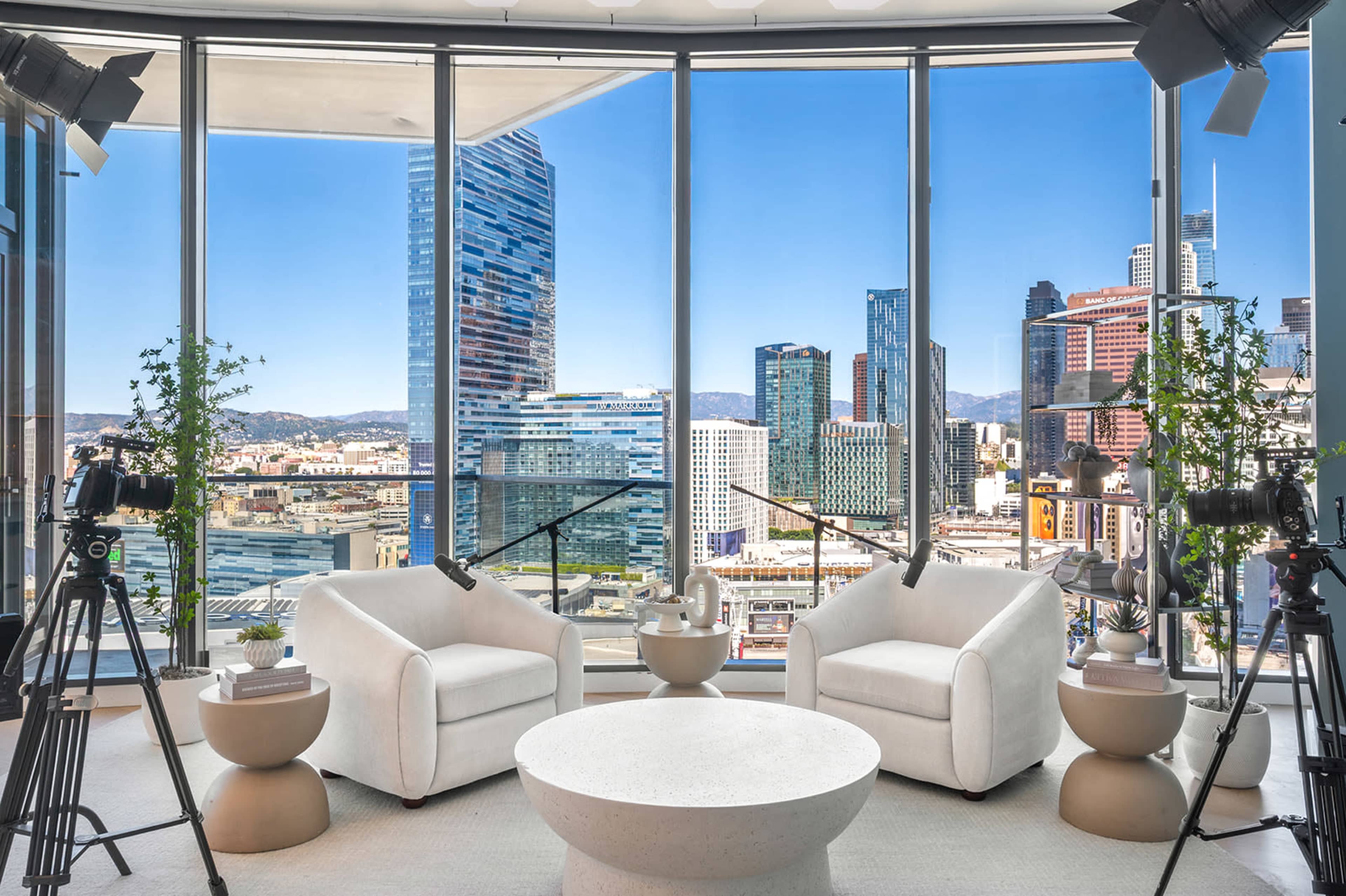 The image shows a modern interview setup with two white chairs and a round table, surrounded by camera equipment, overlooking a city skyline through large windows.