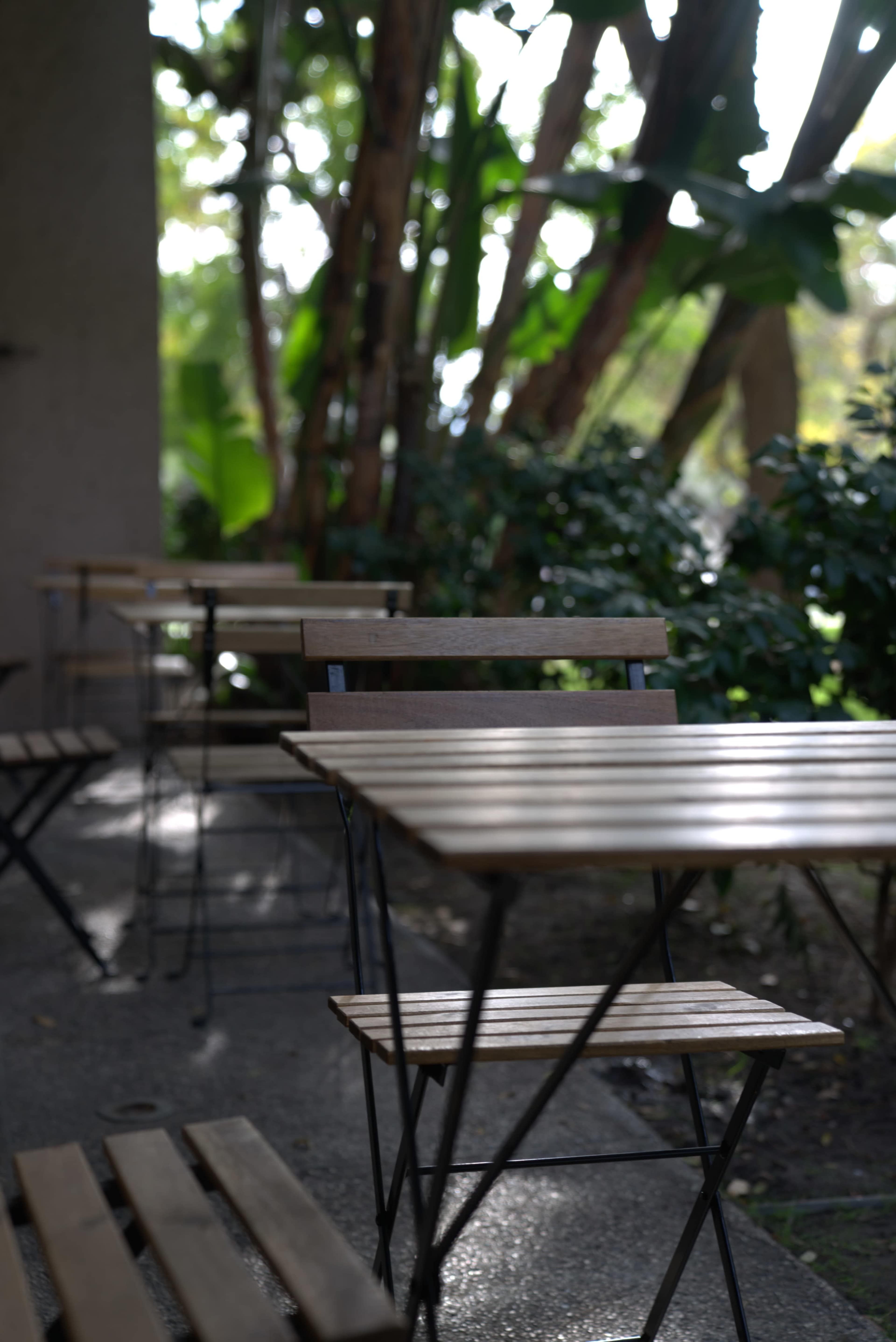 A row of wooden tables with metal frames arranged neatly along a pathway surrounded by greenery.
