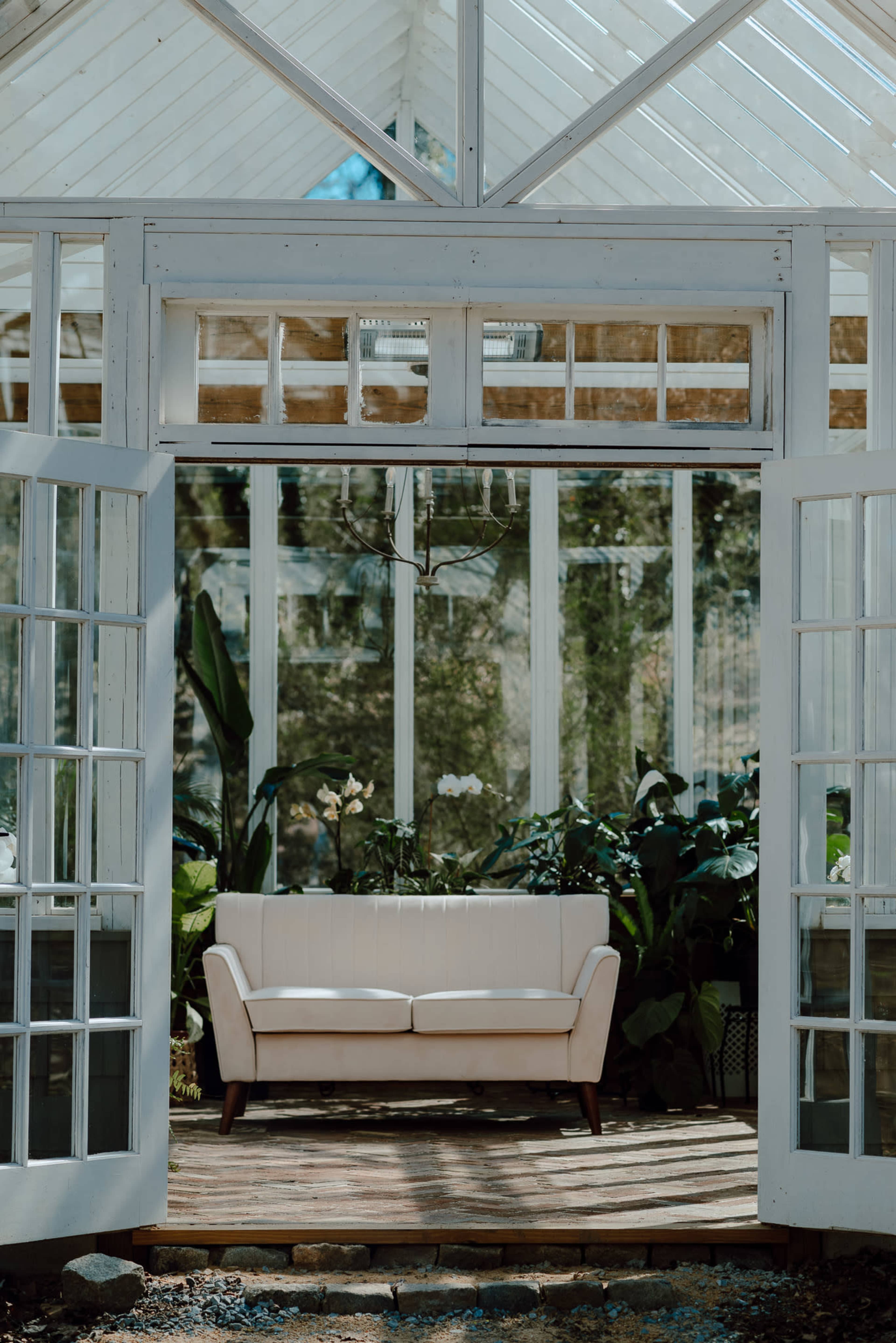 A white sofa sits inside a glass greenhouse surrounded by lush plants.