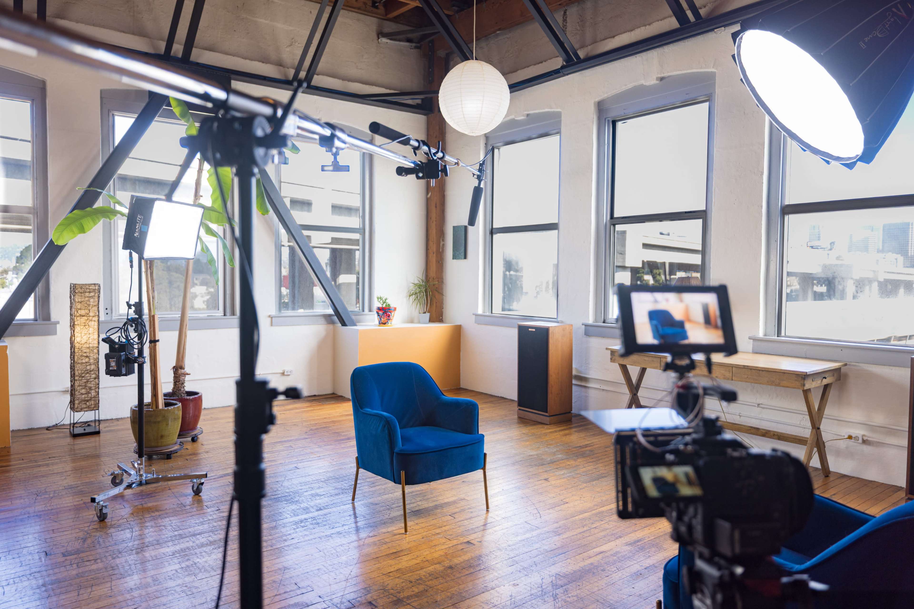 The image features a well-lit filming studio with a blue chair set up in front of a camera and lighting equipment, surrounded by large windows.