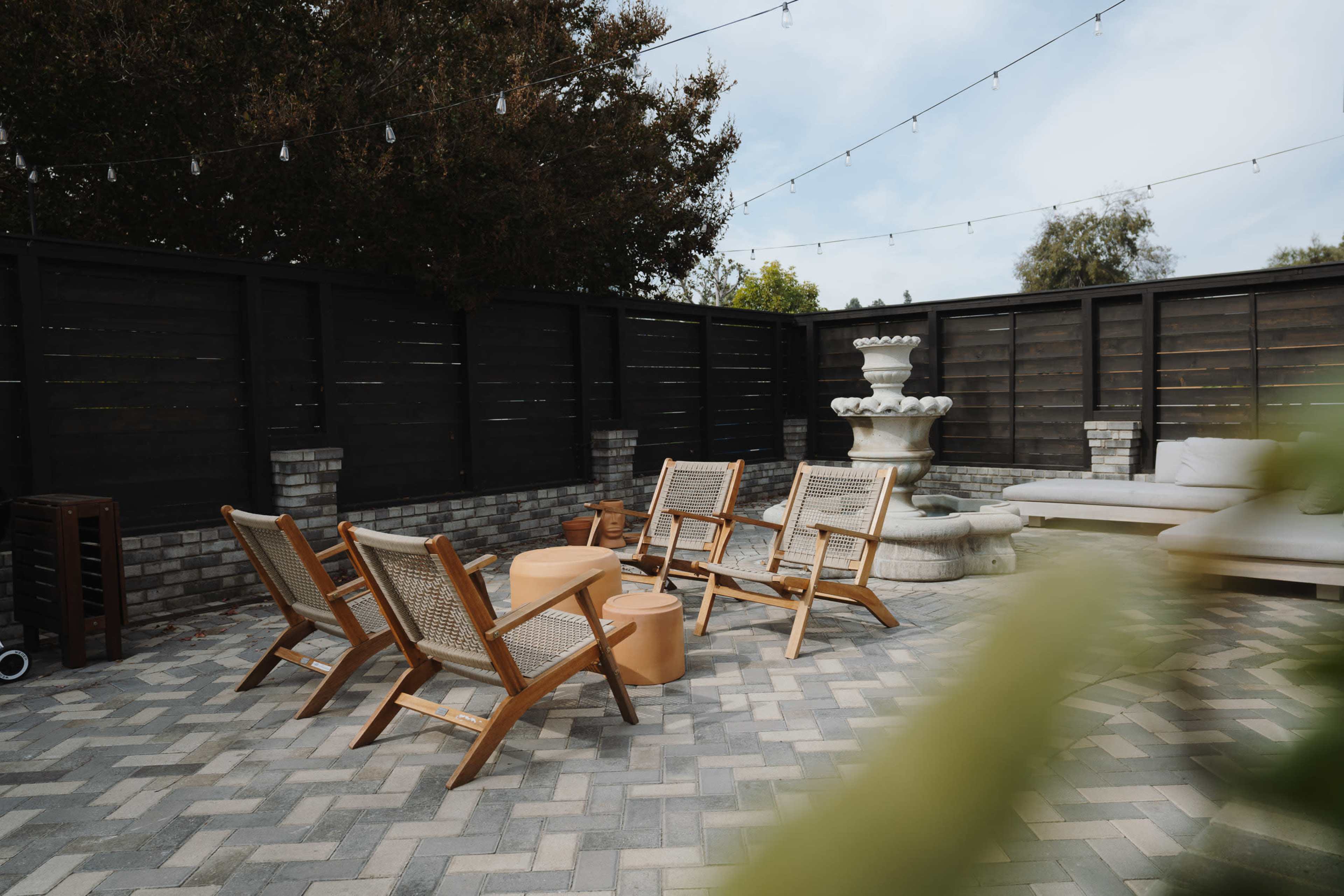 A secluded patio area features wooden lounge chairs arranged around a circular table, with a fountain and a sofa in the background, under string lights.