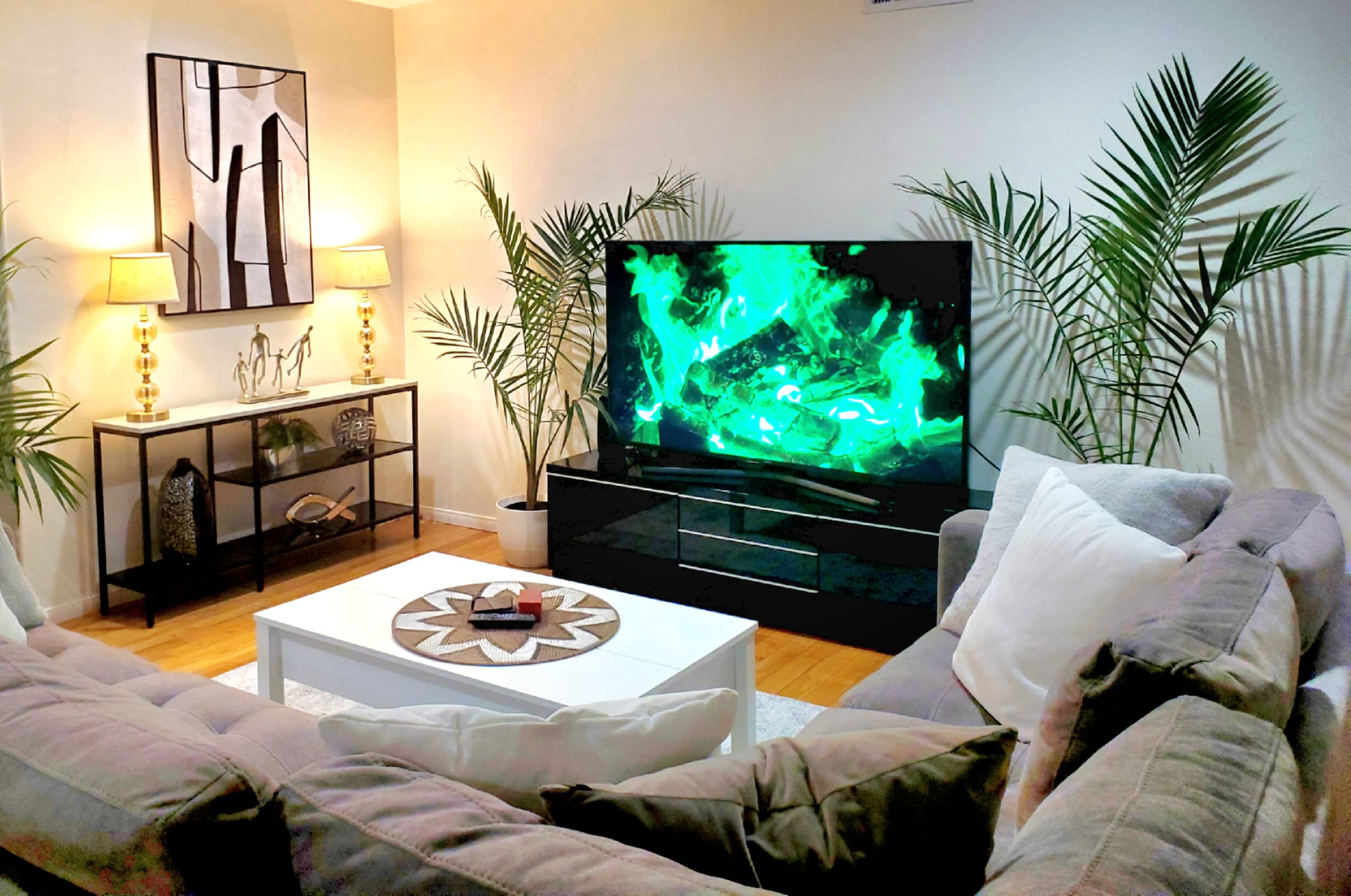 A modern living room with a large TV displaying green visuals, a white coffee table, two lamps, and decorative plants.