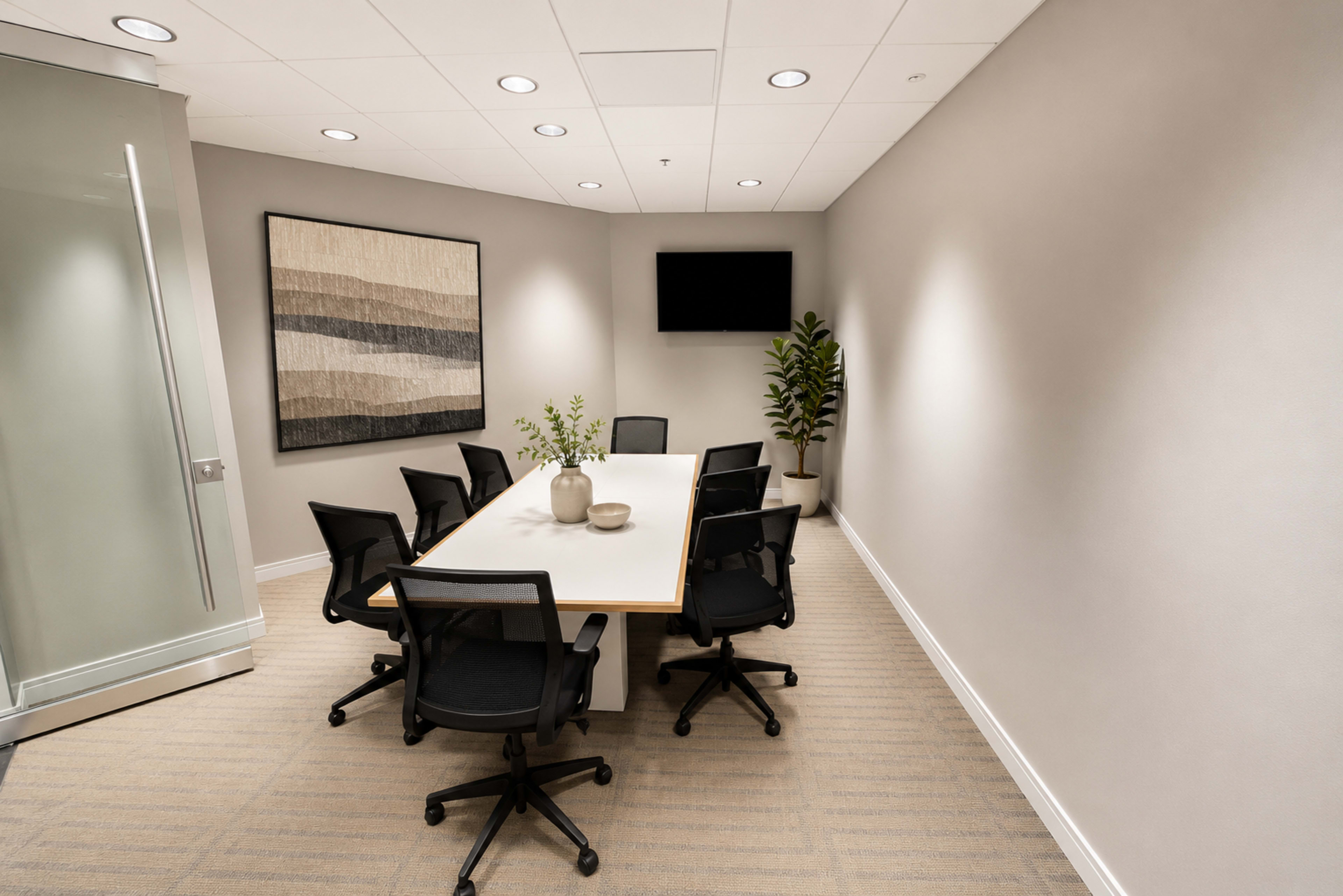 A modern conference room features a rectangular table surrounded by six black chairs, with a plant, artwork, and a wall-mounted television.