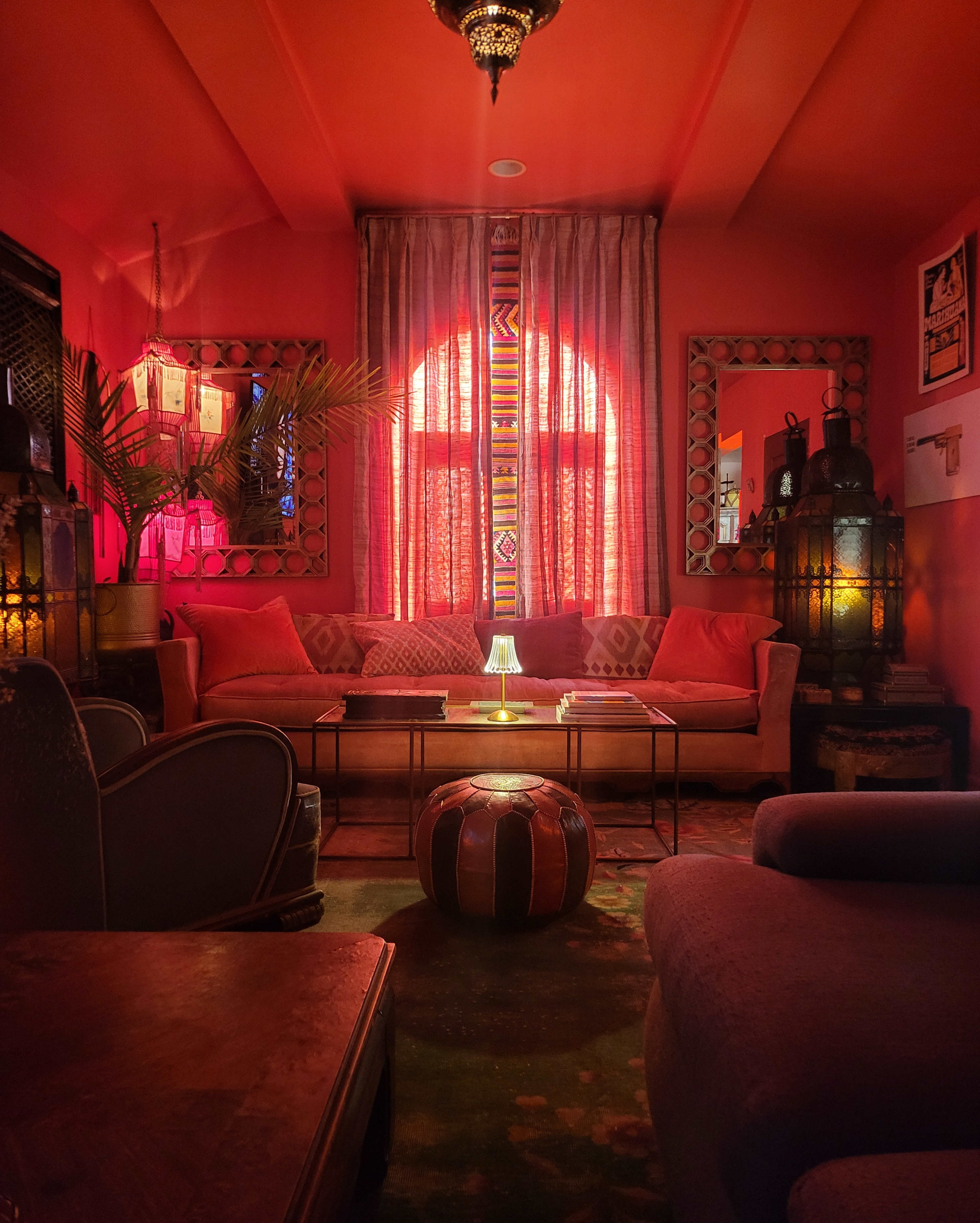 The room features a warm, orange interior with a sofa, decorative cushions, a low table, and lit lanterns against the backdrop of patterned curtains and a large window.