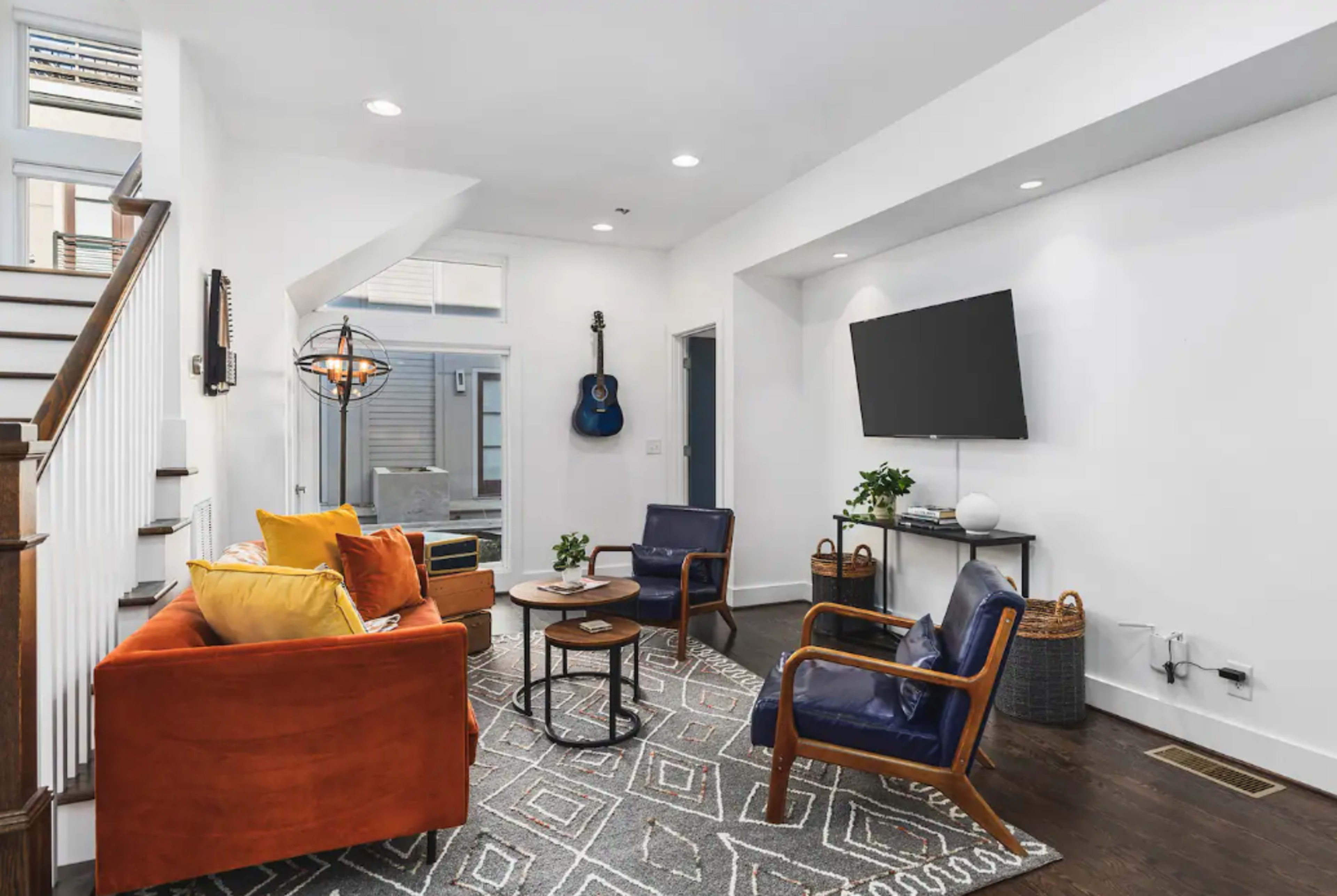 The living room features a mix of orange and blue armchairs, a round coffee table, and a mounted television against a white wall, with a guitar hanging on the adjacent wall.