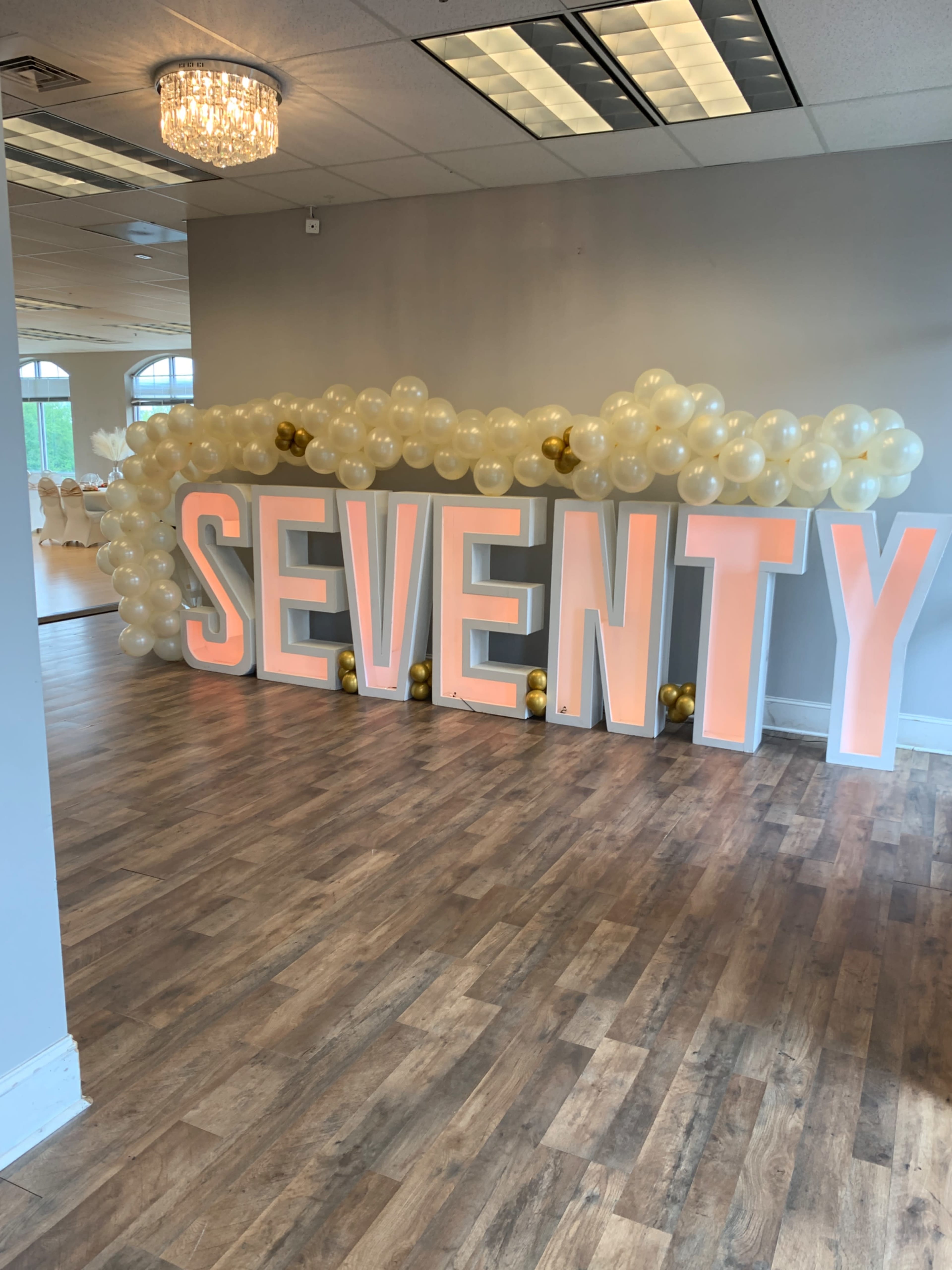 A decorated event space featuring large illuminated letters spelling "SEVENTY" adorned with a backdrop of pale balloons and golden accents.