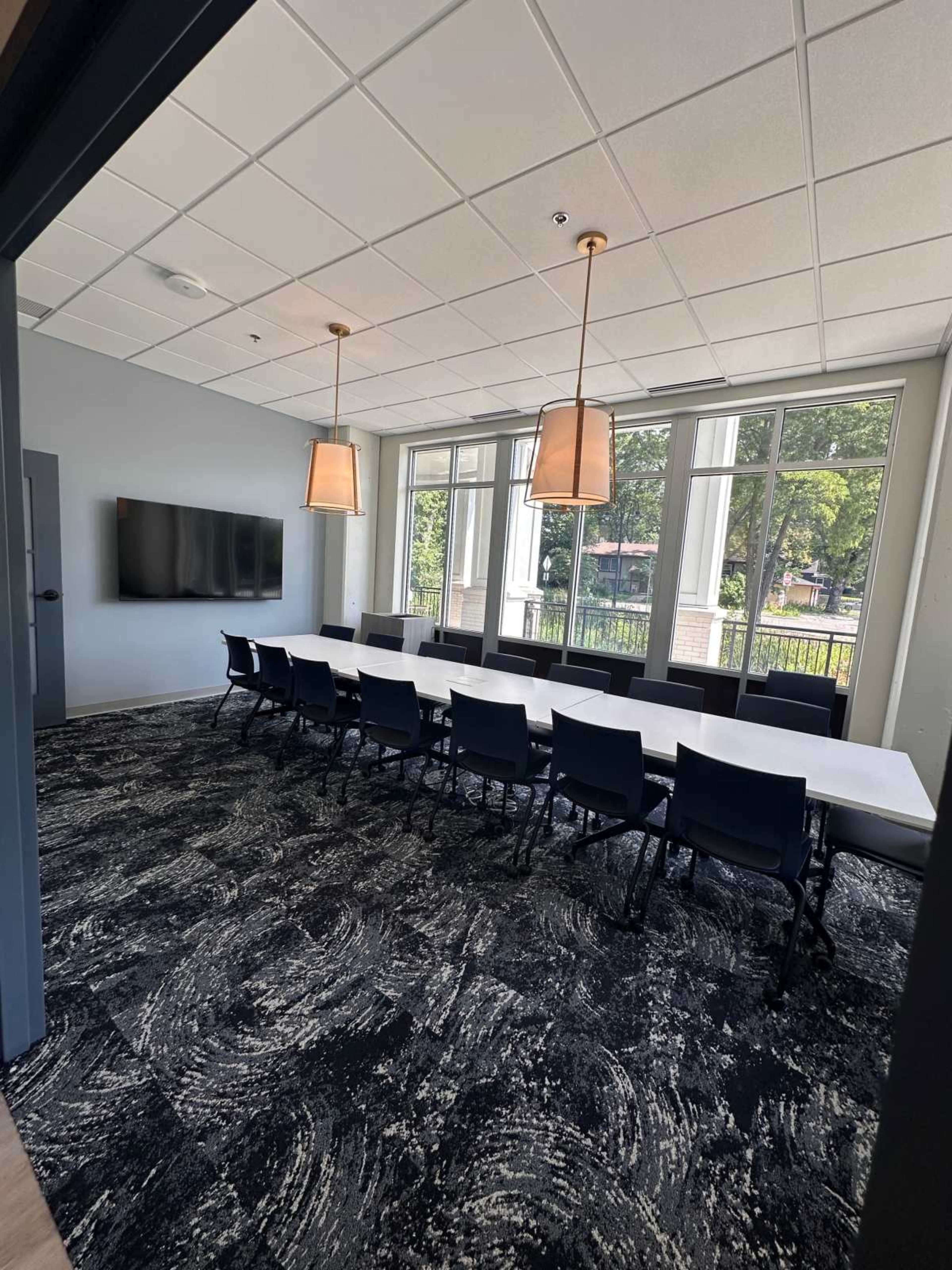 A conference room features a long table surrounded by black chairs, a large screen on the wall, and windows providing natural light.