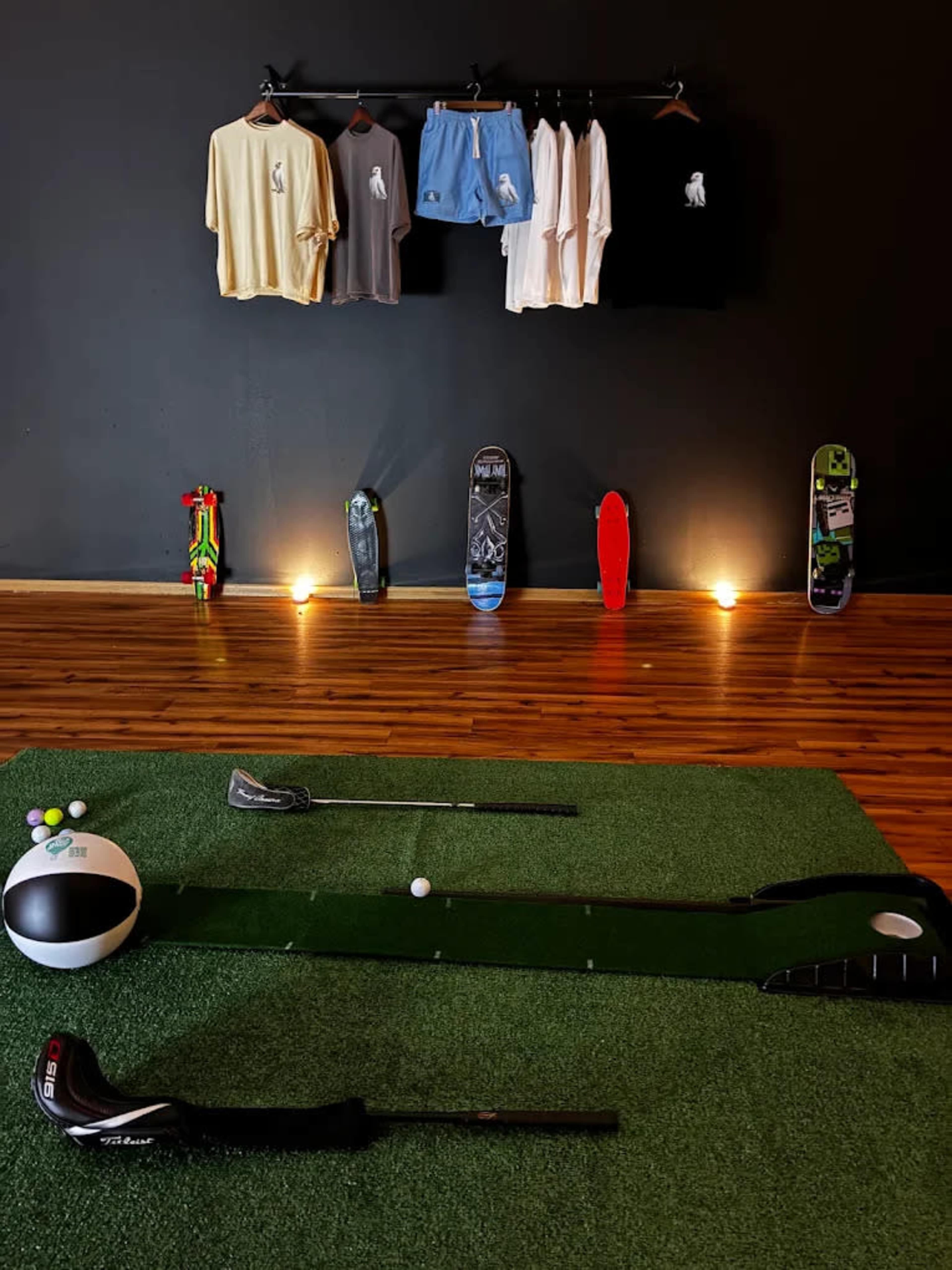 The image shows a display of golf clothing on a rack against a dark wall, with golf equipment arranged on a synthetic green surface in the foreground.