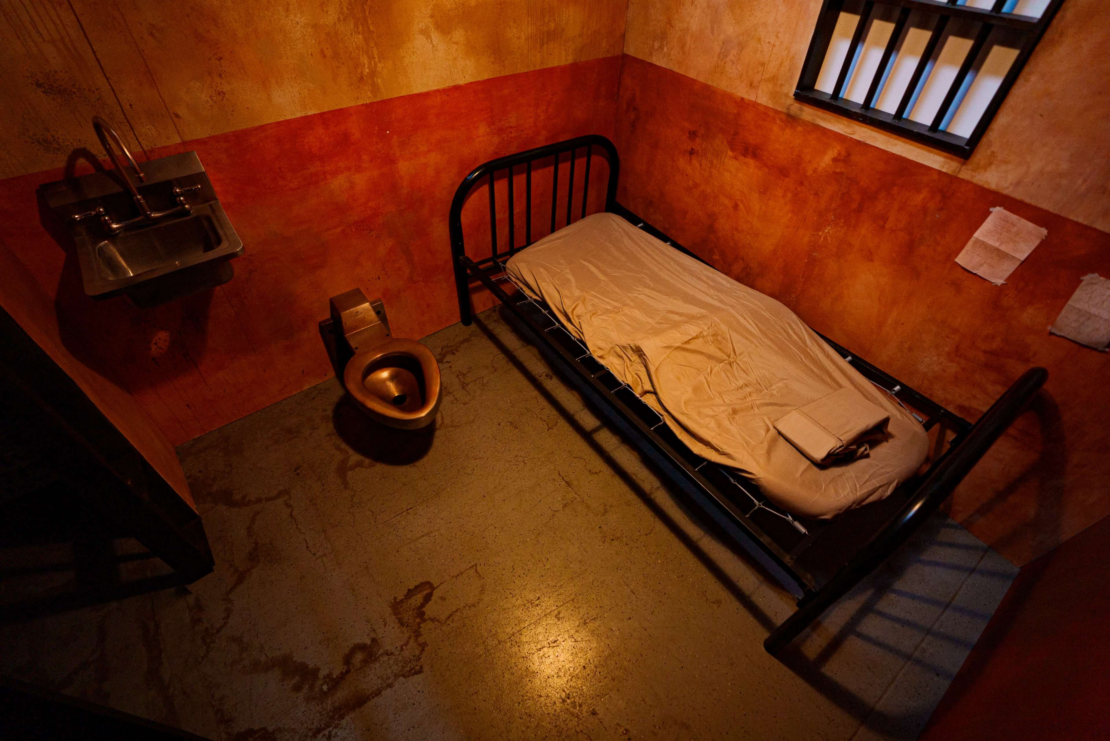 A small, bare prison cell with a metal bed, a toilet, and a sink, all situated against walls painted in shades of brown and orange.