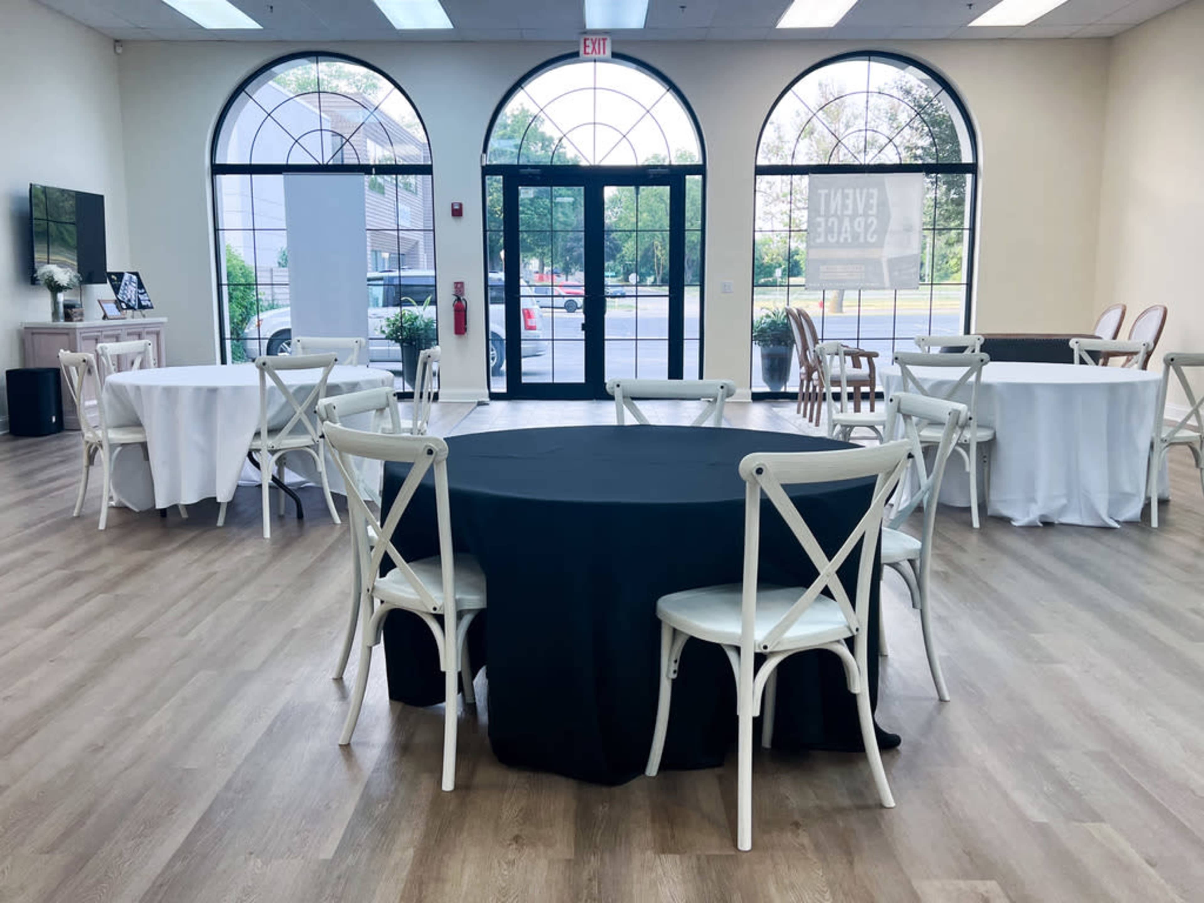 A spacious event room features several round tables with varying tablecloths and large windows allowing natural light.