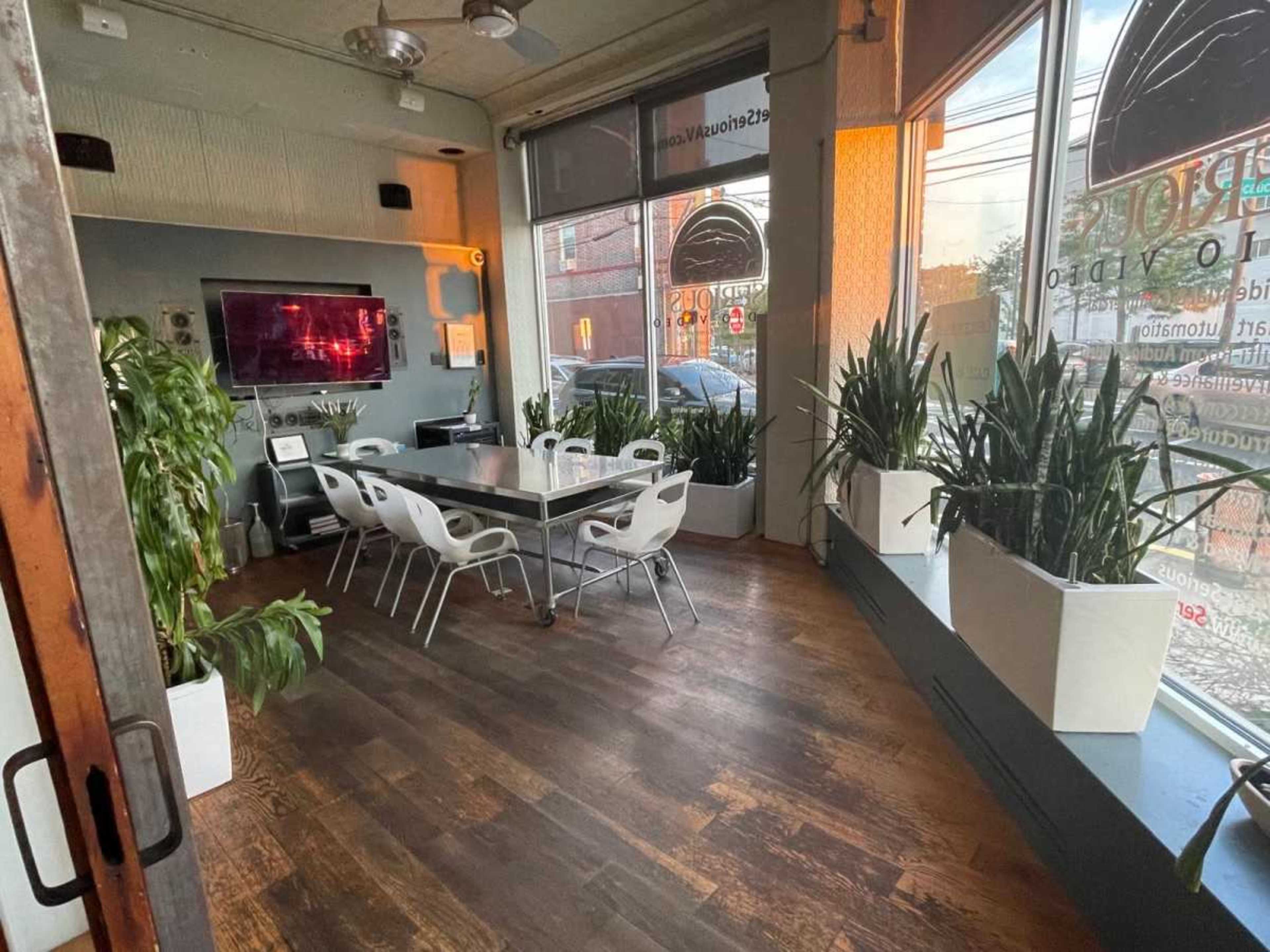 The image shows a modern meeting room with a large table surrounded by white chairs, plants in corner planters, and windows that provide natural light.