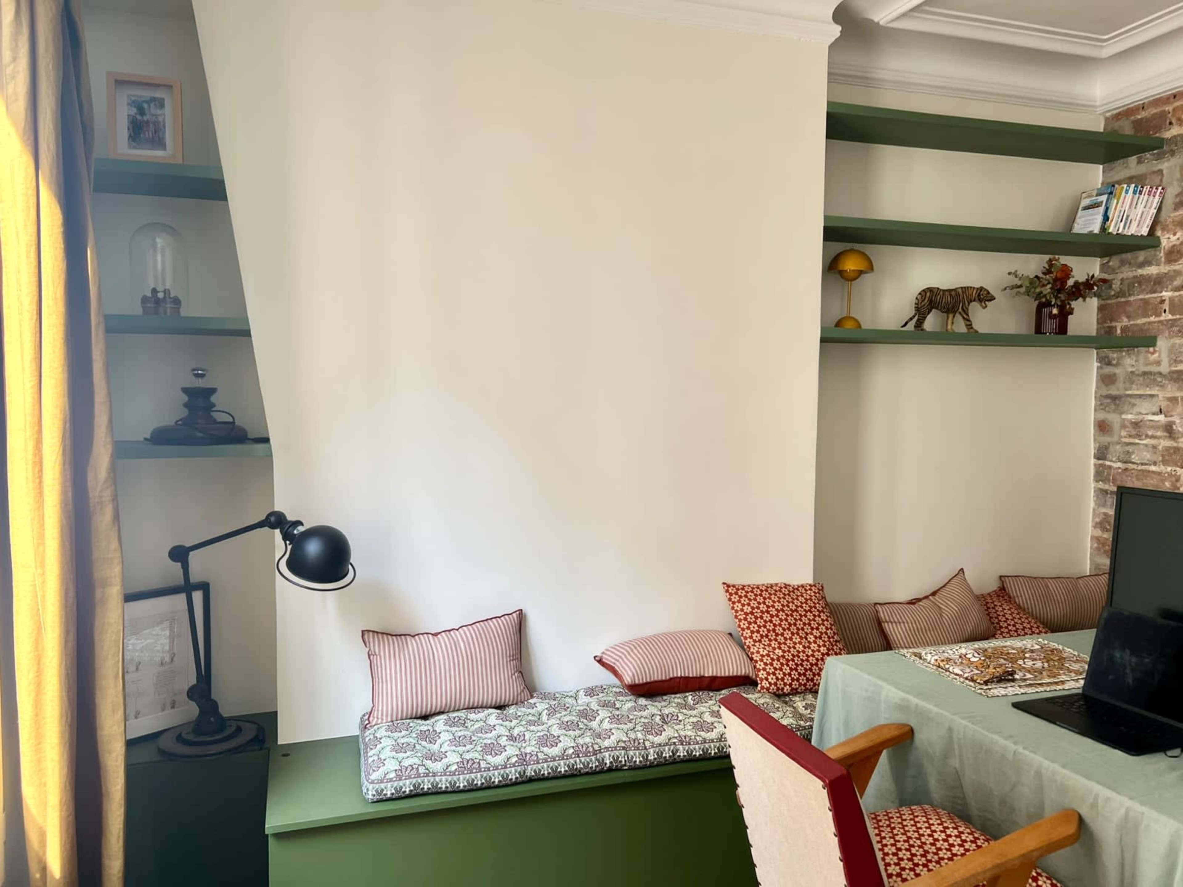 The image shows a corner of a room with a green shelf and a long seating area with cushions, accompanied by a desk featuring a laptop and a chair.