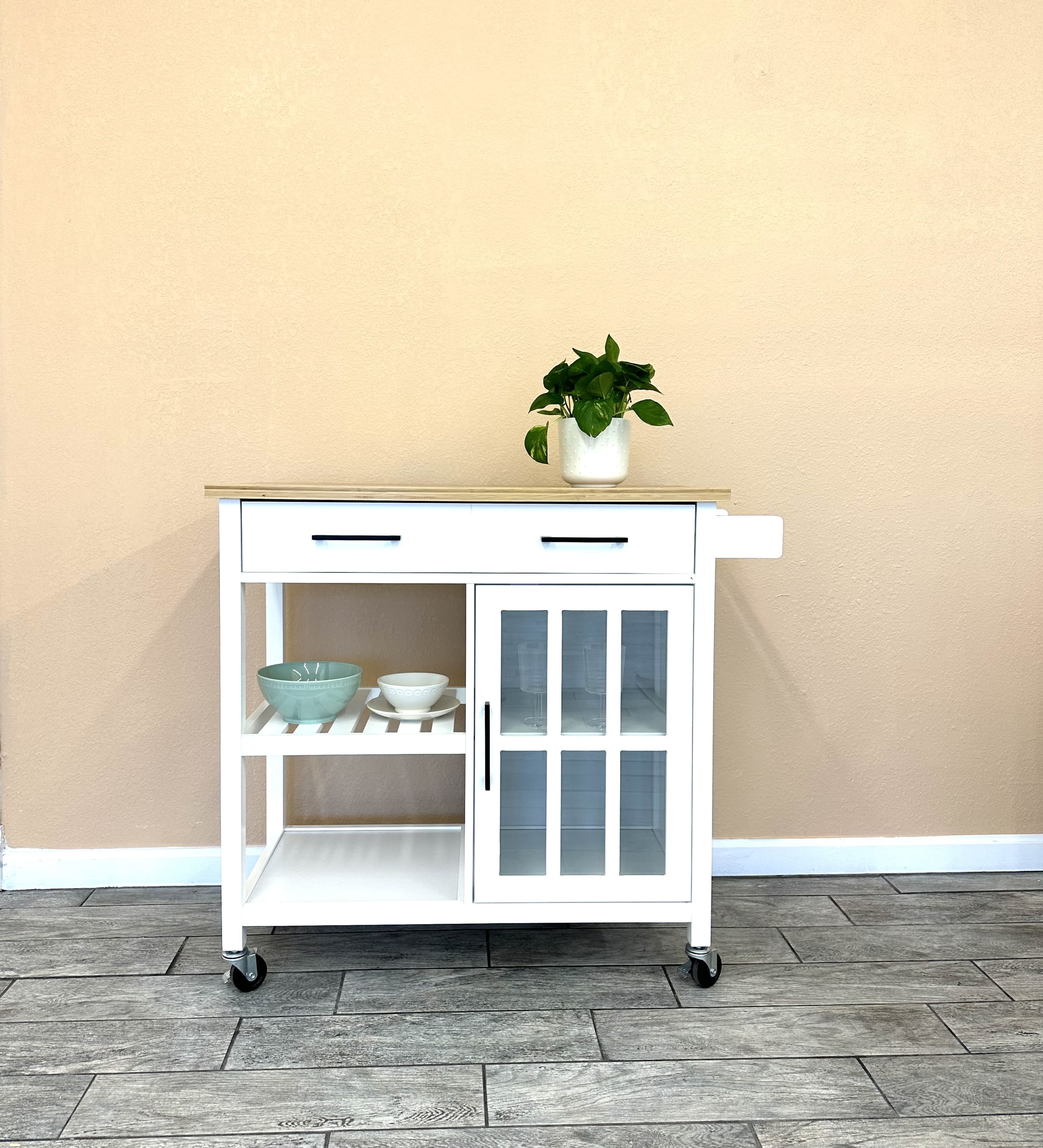 A white kitchen cart with a wooden top, featuring two drawers, a cabinet with glass doors, and open shelving, positioned against a beige wall.
