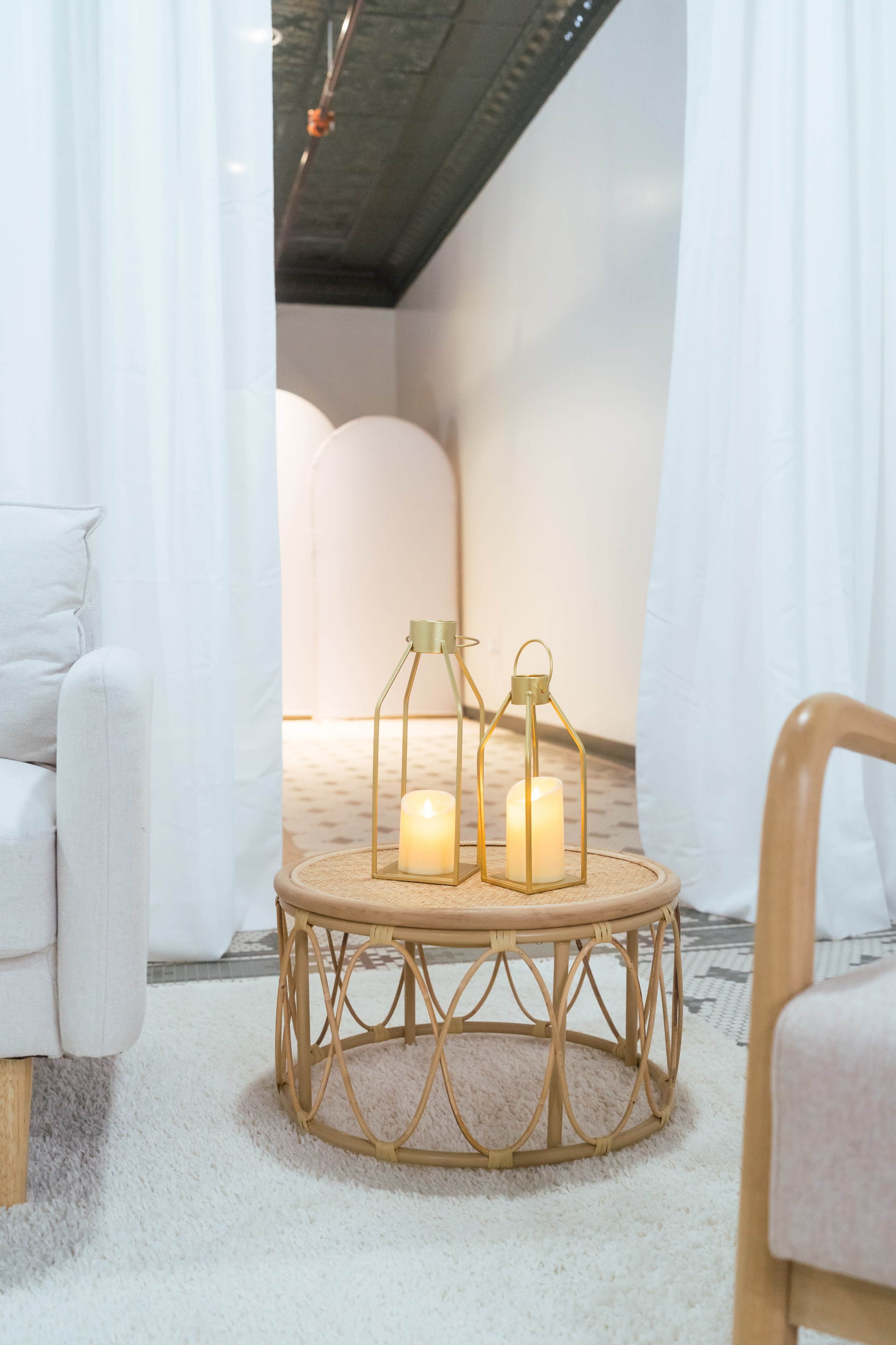 A round woven coffee table with two lanterns and candles is placed on a soft rug, in a room with white curtains and a partially visible backdrop.