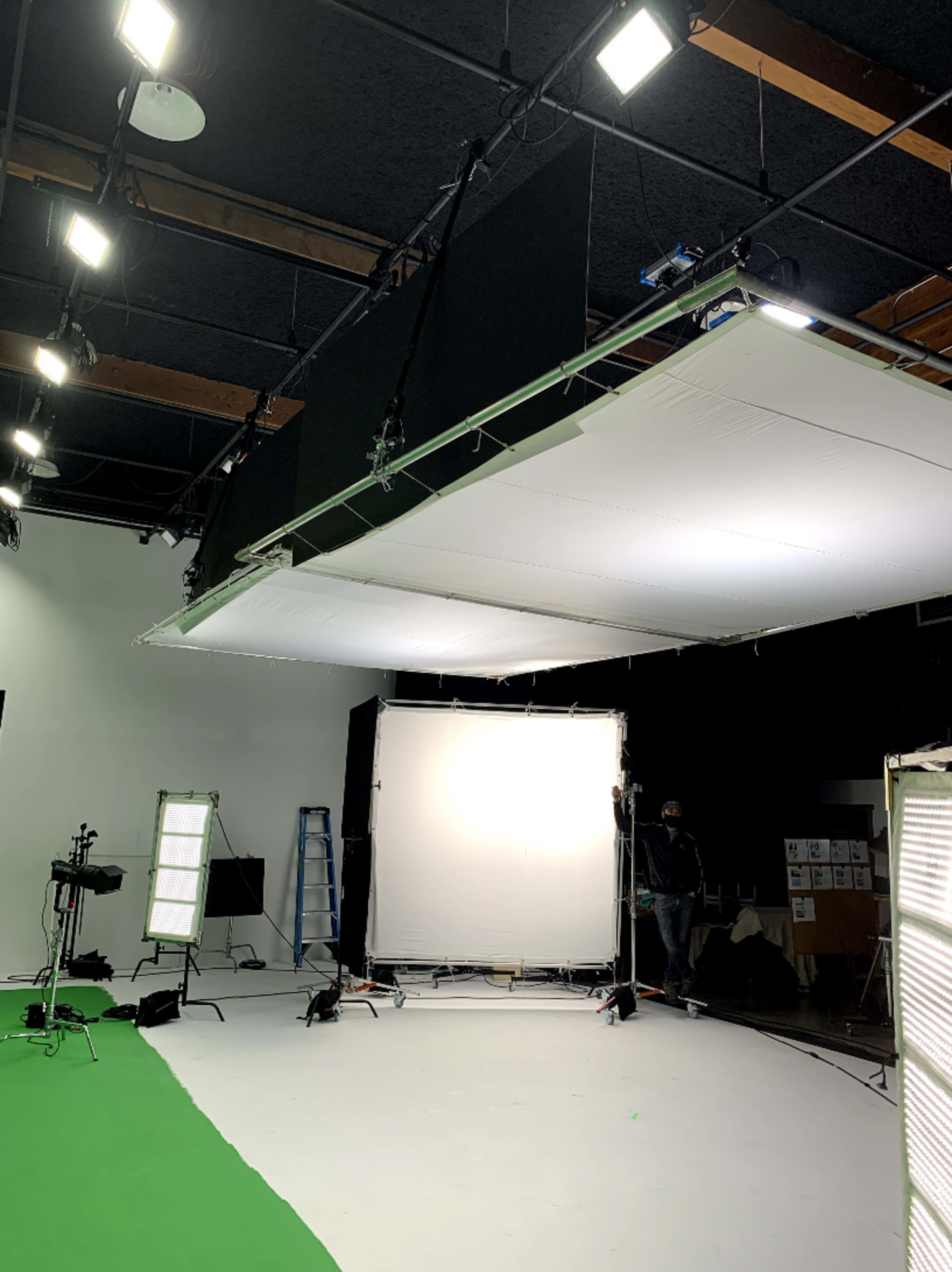 The image shows a film studio setup with lighting equipment, a green screen, and several large white backdrops.