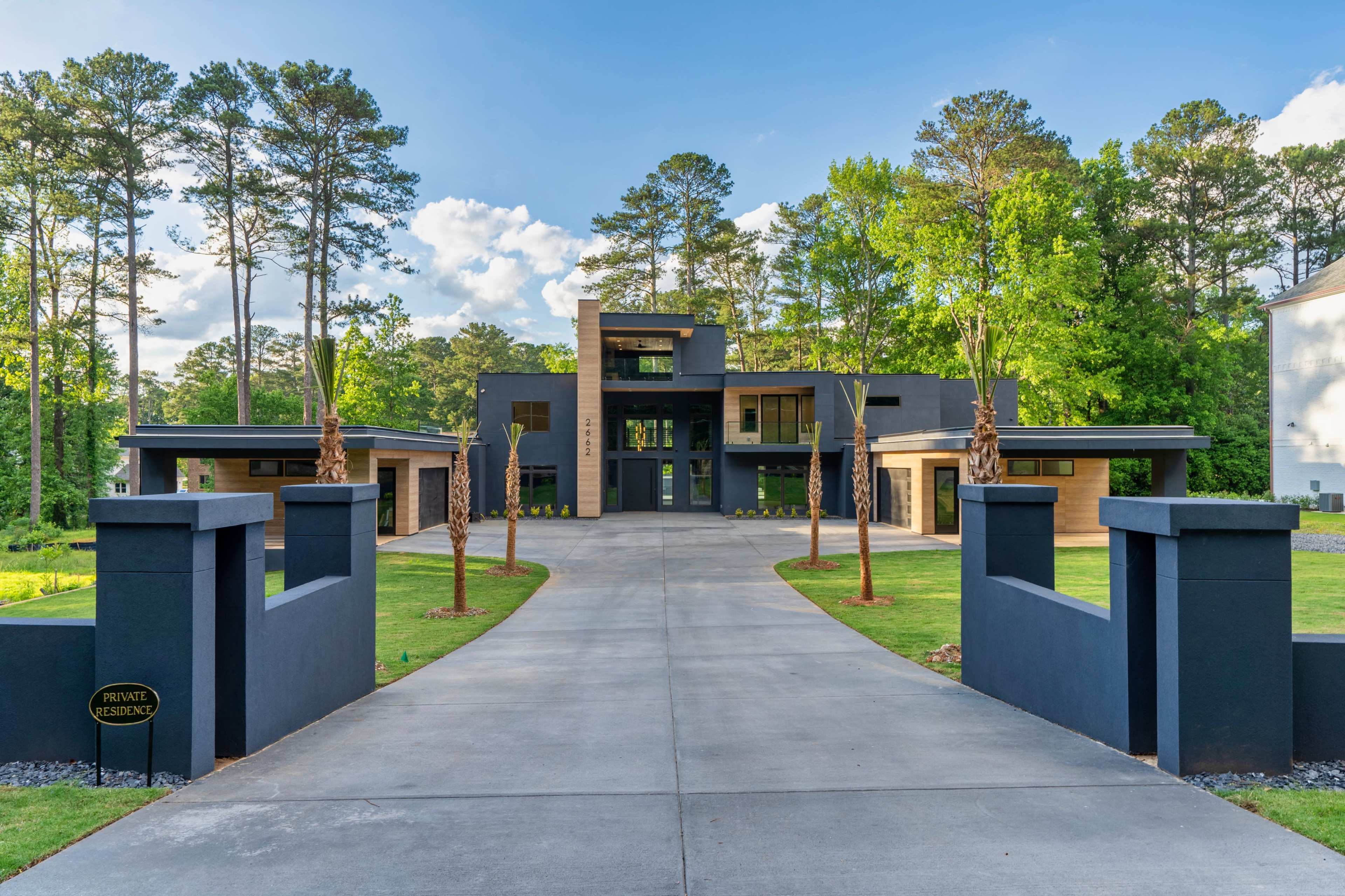 A modern house with a symmetrical design and a long driveway leading to its entrance, surrounded by tall trees.