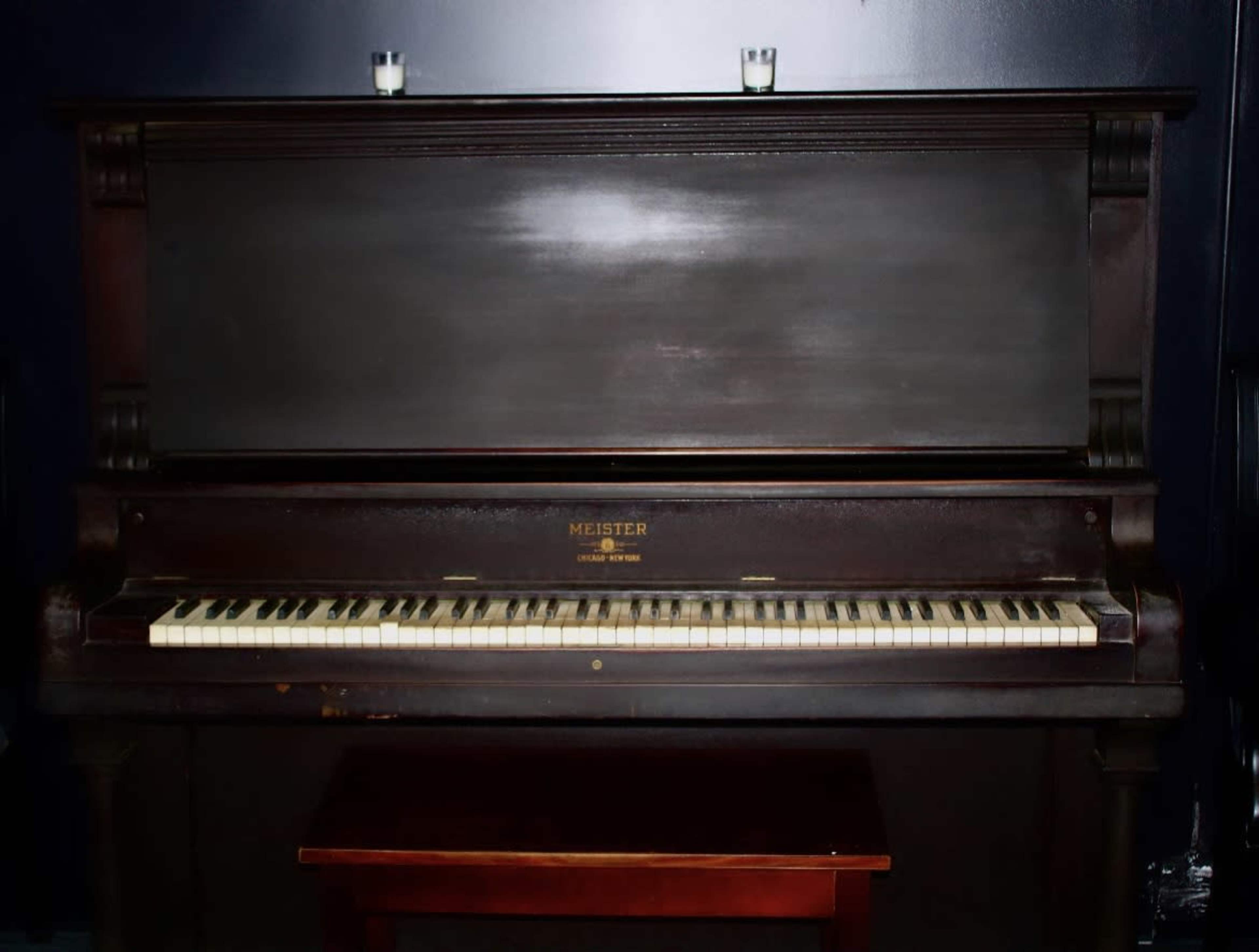 A dark wooden upright piano stands against a wall, with two candles on top and a small table in front.